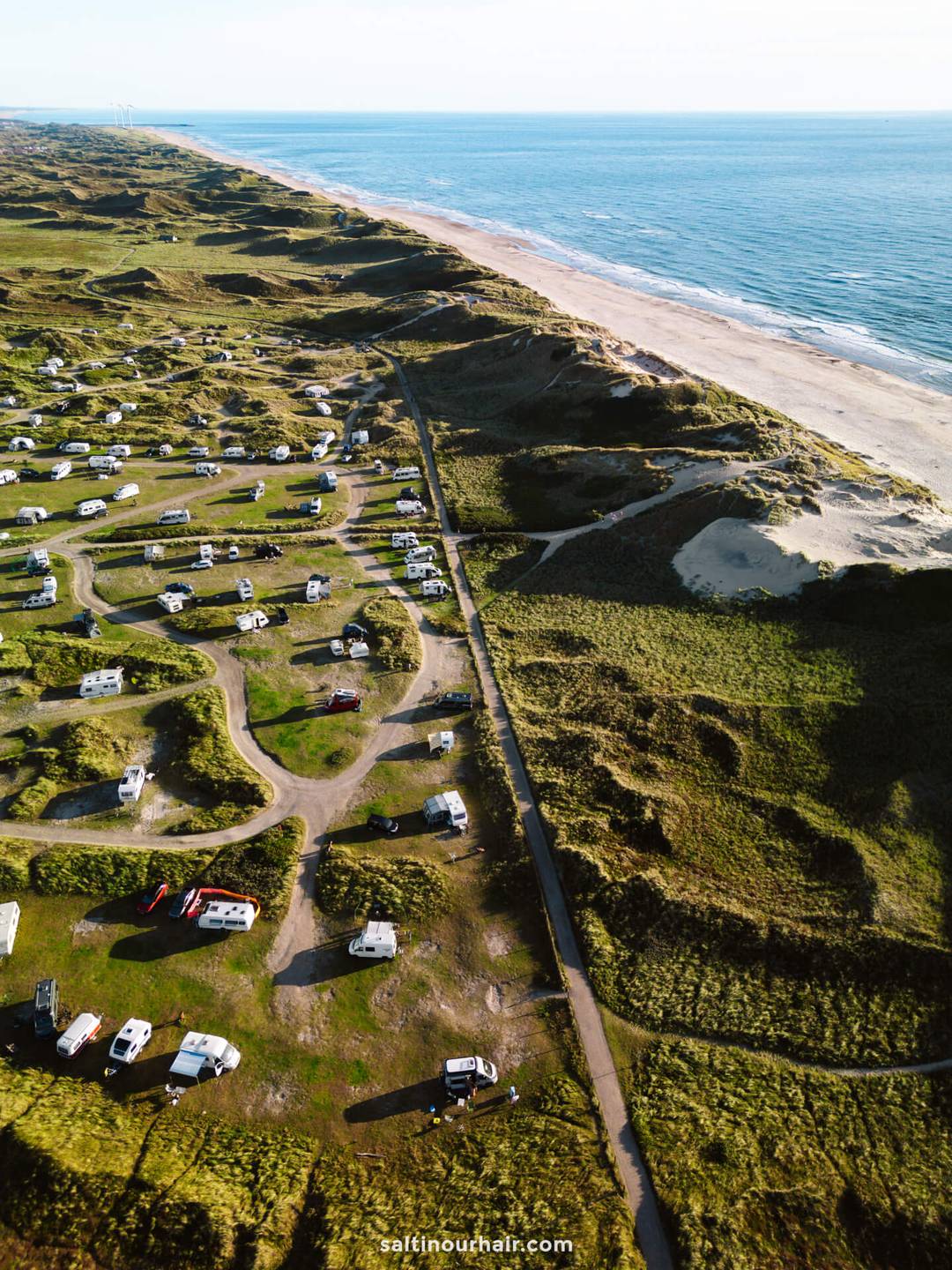 Aerial view of a coastal campsite, perfect for any Denmark itinerary, with numerous RVs and winding roads near dunes and a sandy beach beside the ocean.