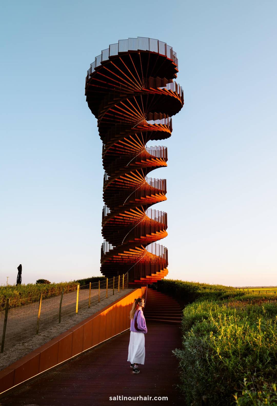 A person stands at the base of a tall, spiral observation tower called Marsk Tower at sunset&mdash;an unforgettable moment for any Denmark itinerary&mdash;as another individual takes in the view from the top, all surrounded by lush greenery and an open sky.