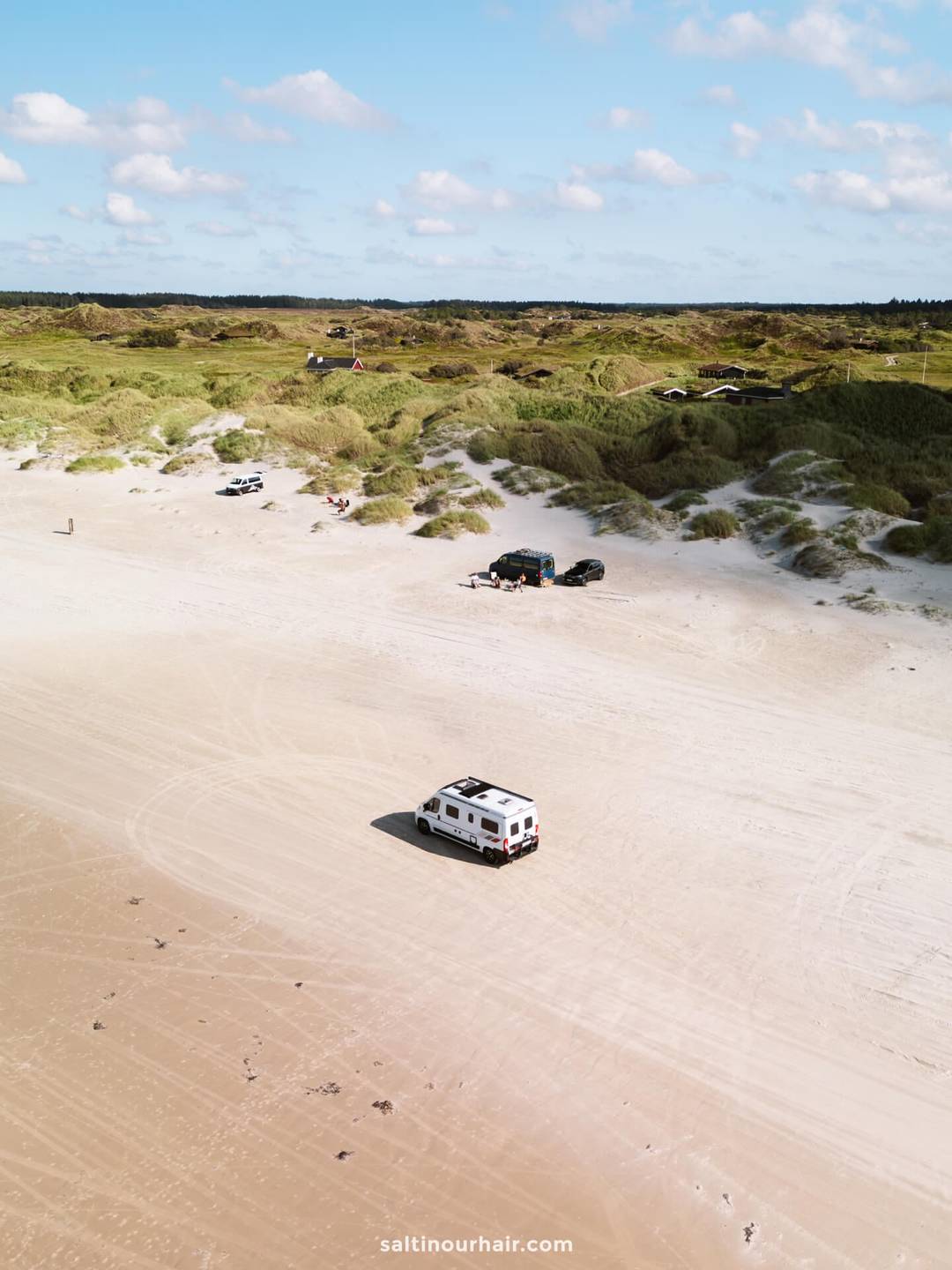 A white camper van drives on a wide sandy beach is the perfect addition to any Denmark road trip itinerary, with several vehicles and grassy dunes in the background under a partly cloudy sky.