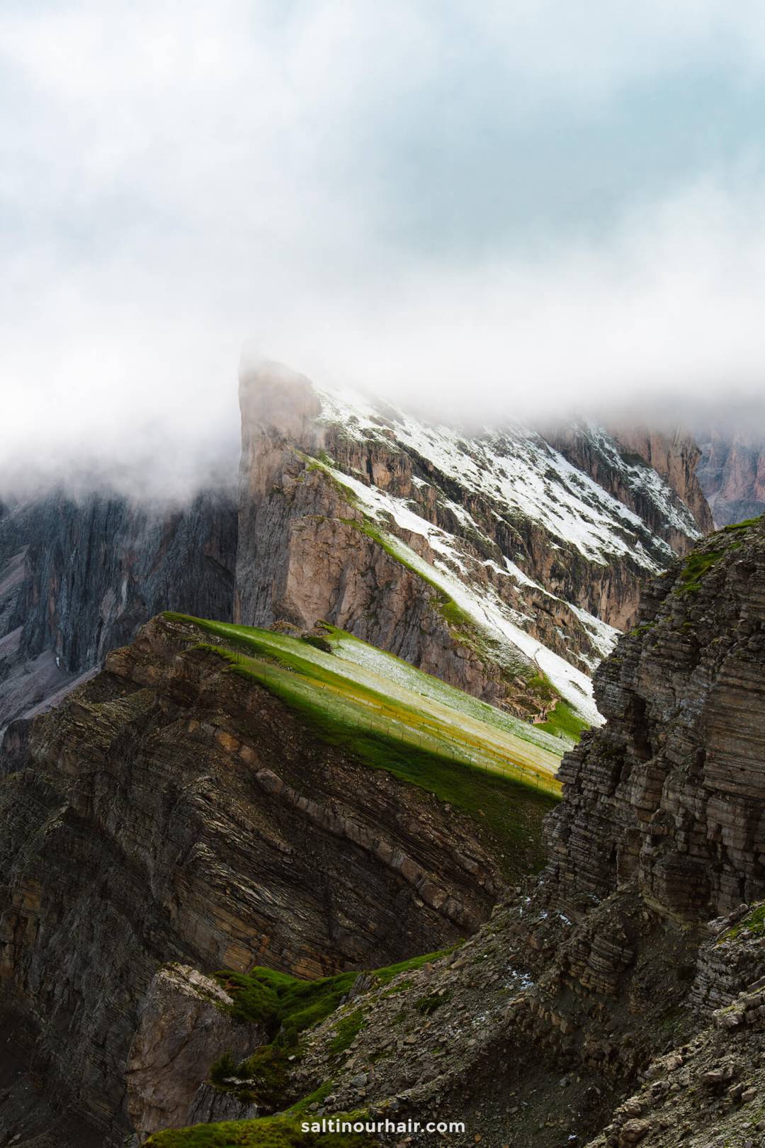 Een rotsachtig berglandschap met stukken groen gras en sneeuw, gedeeltelijk bedekt door laaghangende wolken, omlijst de dramatische Seceda bergkam in de Dolomieten van Itali&euml;