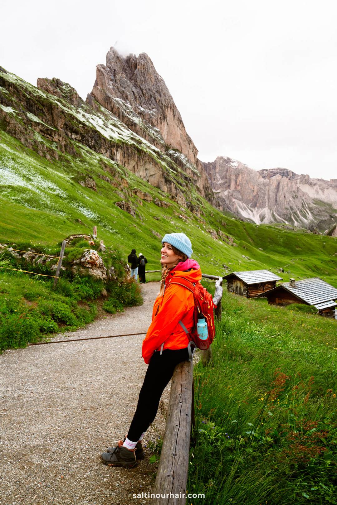 Een vrouw in een rode jas en blauwe muts zit op een houten hek langs een bergpad, met de prachtige Seceda bergkam en rotsachtige pieken die oprijzen achter groene heuvels van de Dolomieten Itali&euml;