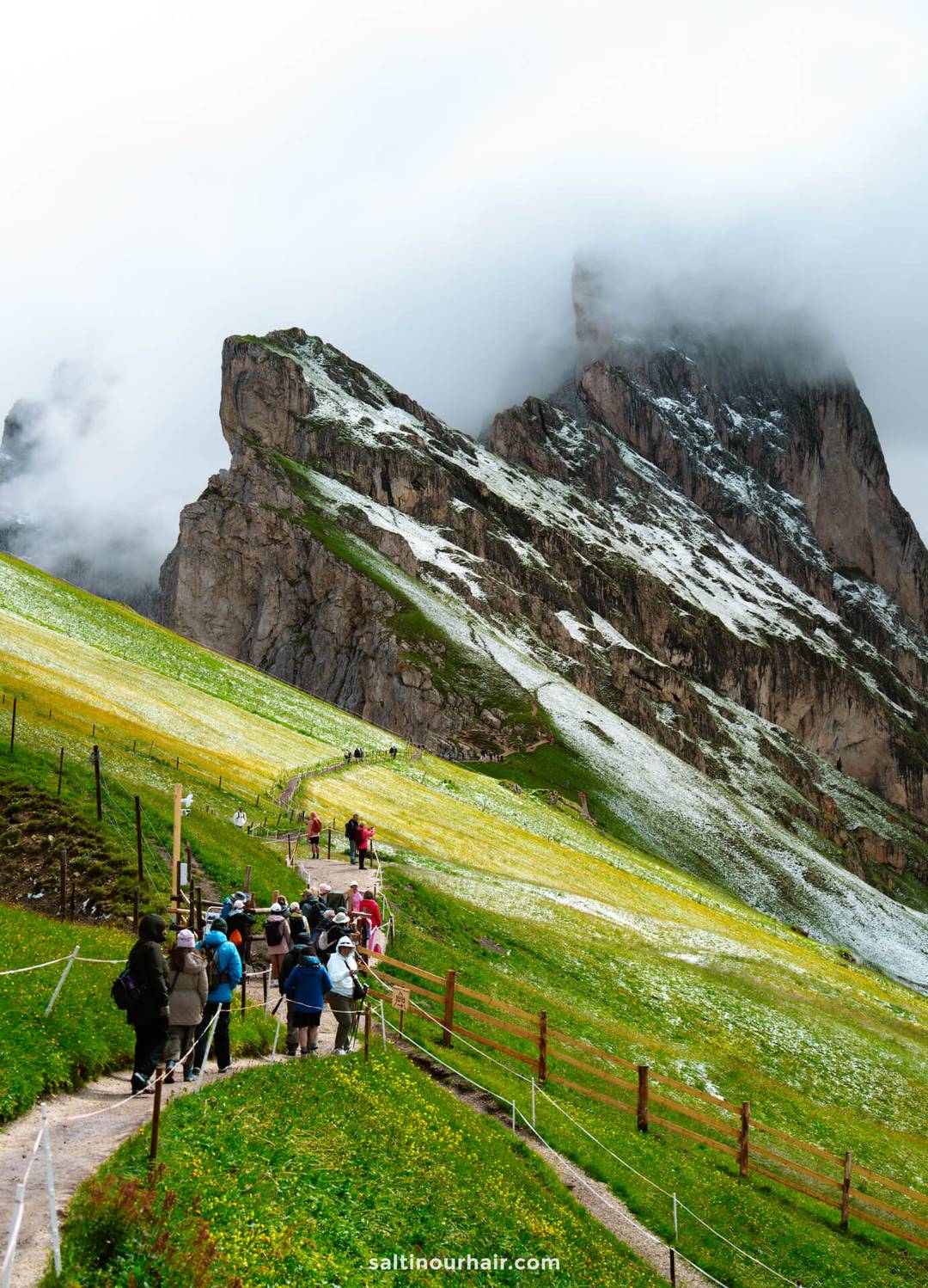 Groep wandelaars loopt over een slingerend pad op een groene heuvelflank met sneeuwvlekken, richting de dramatische Seceda-bergkam en steile, gedeeltelijk door wolken bedekte rotspieken.