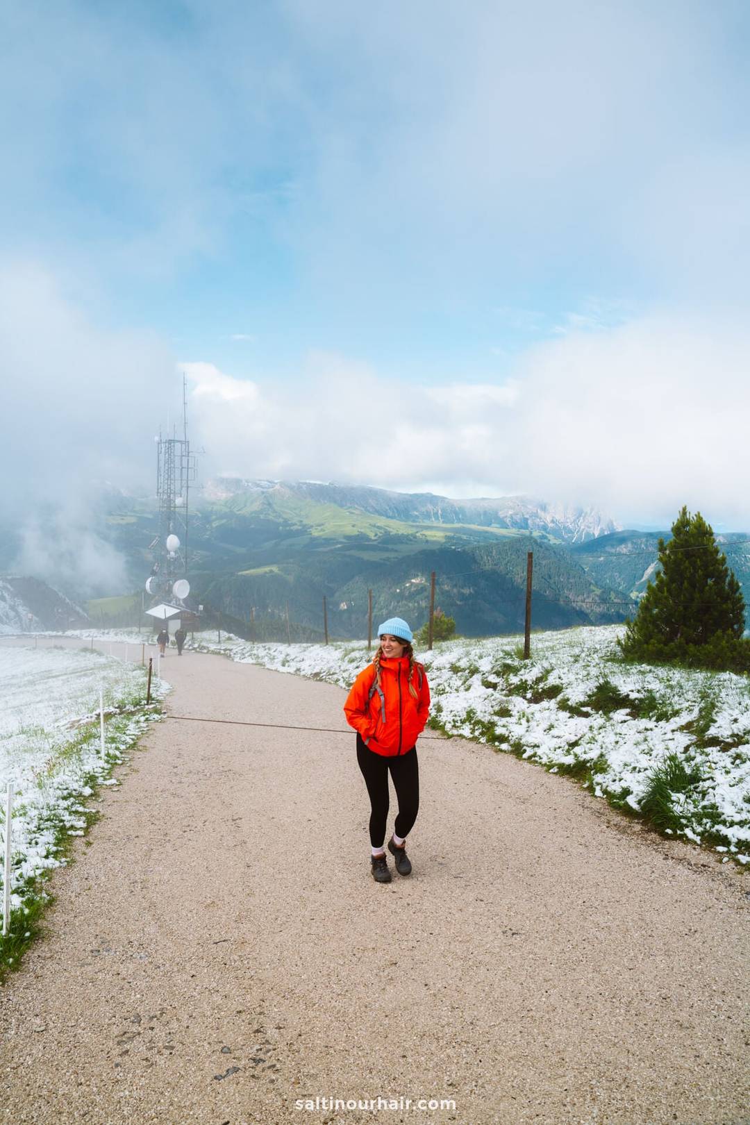 Persoon in rood jack en blauwe helm staat op een grindpad langs de Seceda-bergkam in een bergachtig gebied met sneeuwvlekken en groen onder een bewolkte lucht.