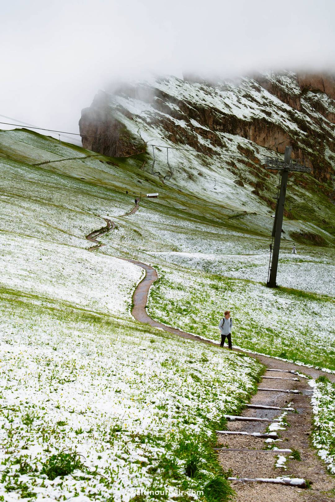 Persoon loopt omhoog over een slingerend pad door een grasrijke, licht besneeuwde heuvelflank bij de Seceda-bergkam, met hoogspanningsleidingen en lage wolken boven een rotsachtige berg.