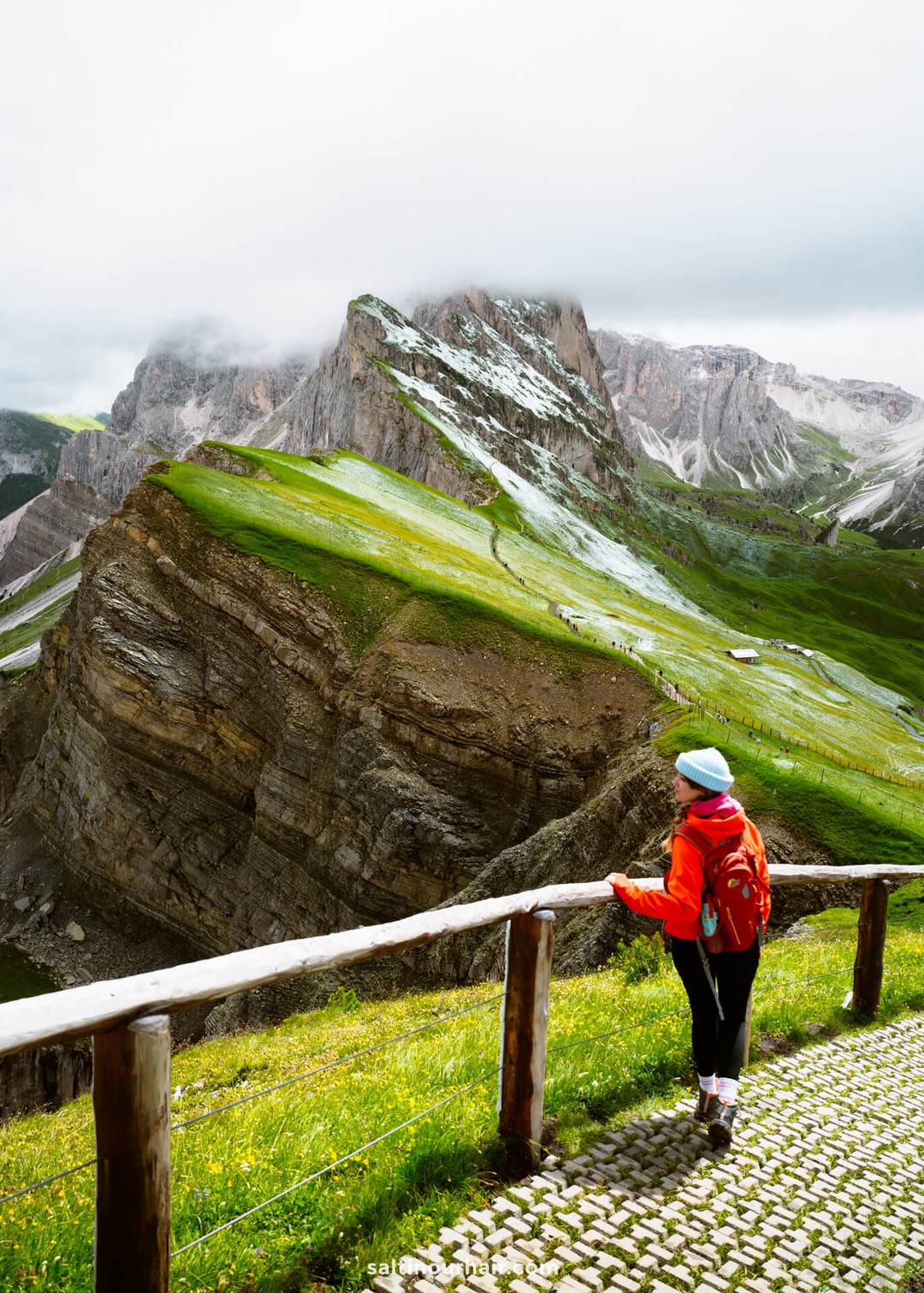 Persoon in rood jack en blauwe muts staat bij een houten leuning en kijkt uit over de dramatische groene en rotsachtige Seceda-bergkam, gedeeltelijk gehuld in wolken.