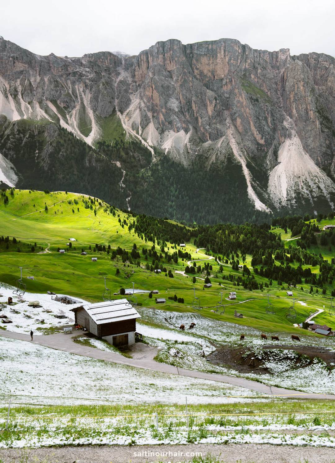 Een rustieke schuur staat op een besneeuwd stuk tussen groene velden bij de Seceda bergkam in de Dolomieten, met verspreide huizen en dramatische bergen op de achtergrond onder een bewolkte hemel.