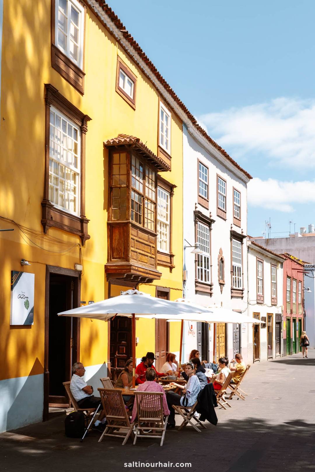 Mensen zitten aan tafeltjes onder parasols buiten een caf&eacute; in een zonnige straat in San Crist&oacute;bal de La Laguna, Tenerife, omringd door kleurrijke, traditionele gebouwen in Europese stijl.