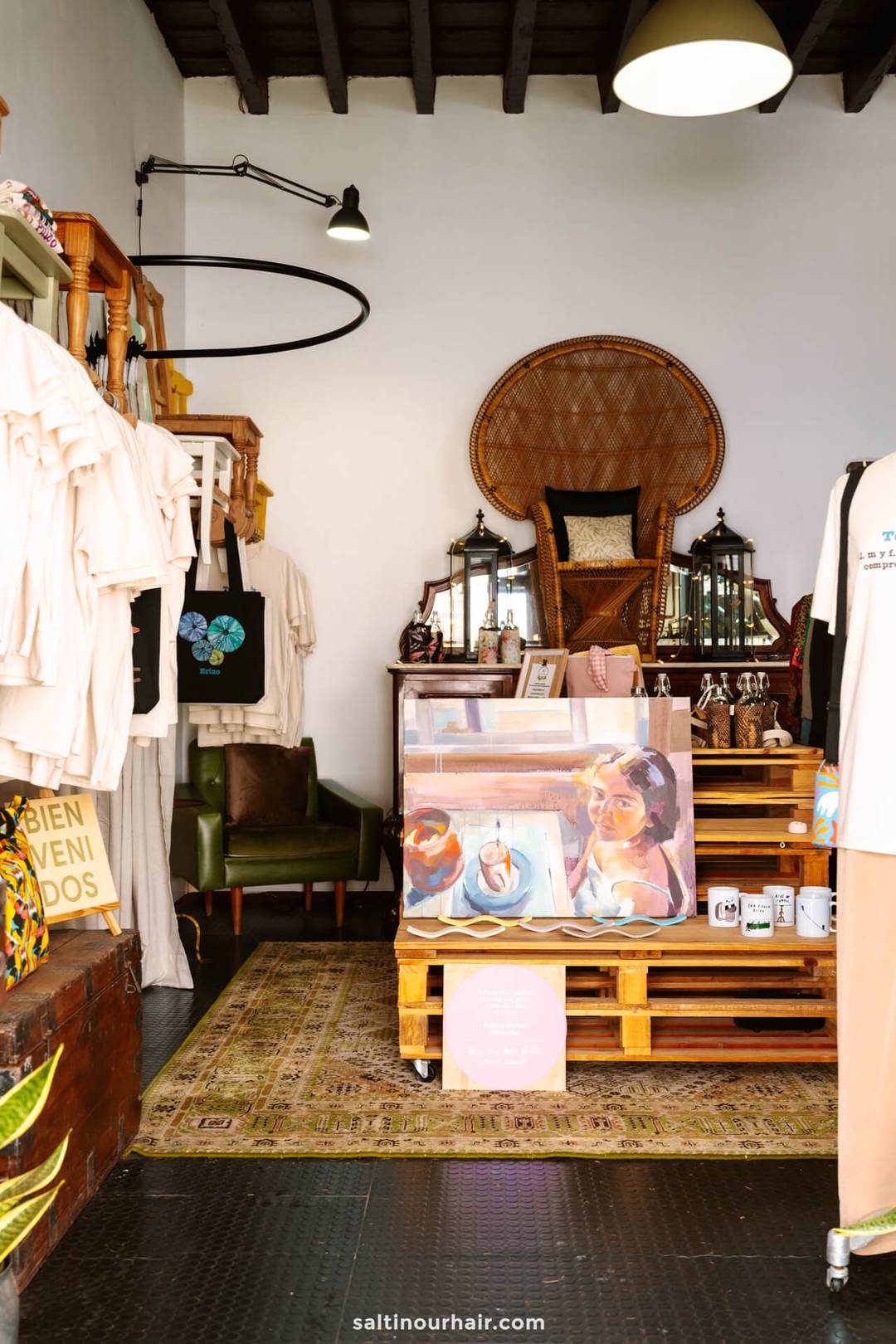 Een kleine boetiek in San Crist&oacute;bal de La Laguna, Tenerife, toont kleding, draagtassen, kunstprints, mokken en decoratieve lantaarns, met een grote rieten stoel en diverse items op houten pallets en planken.