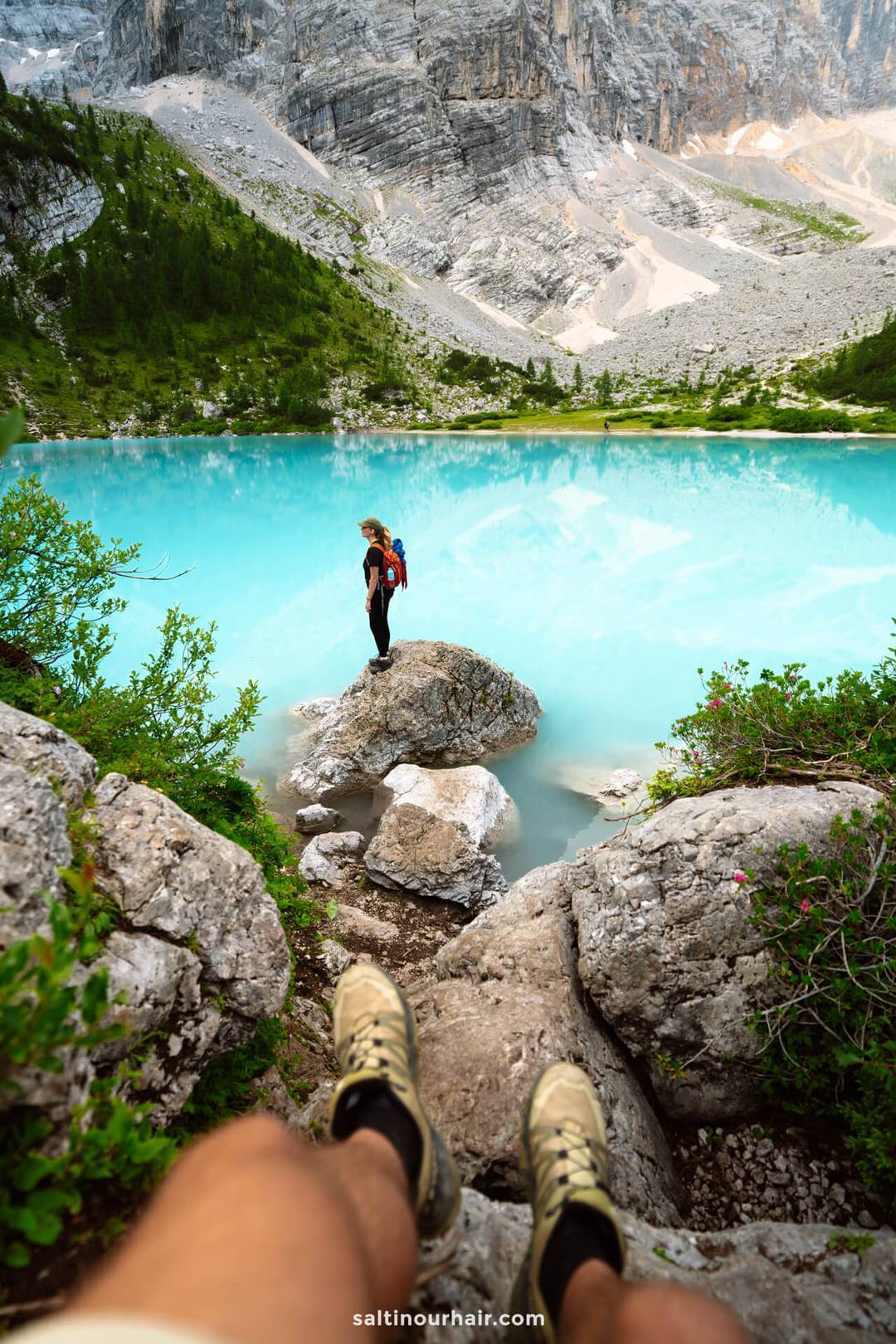 Persoon met rugzak staat op een rots bij een turquoiseblauw meer omgeven door bergen, gezien vanuit het perspectief van iemand anders die zit met benen op de voorgrond, een inspirerend moment voor elke Dolomieten reisroute.