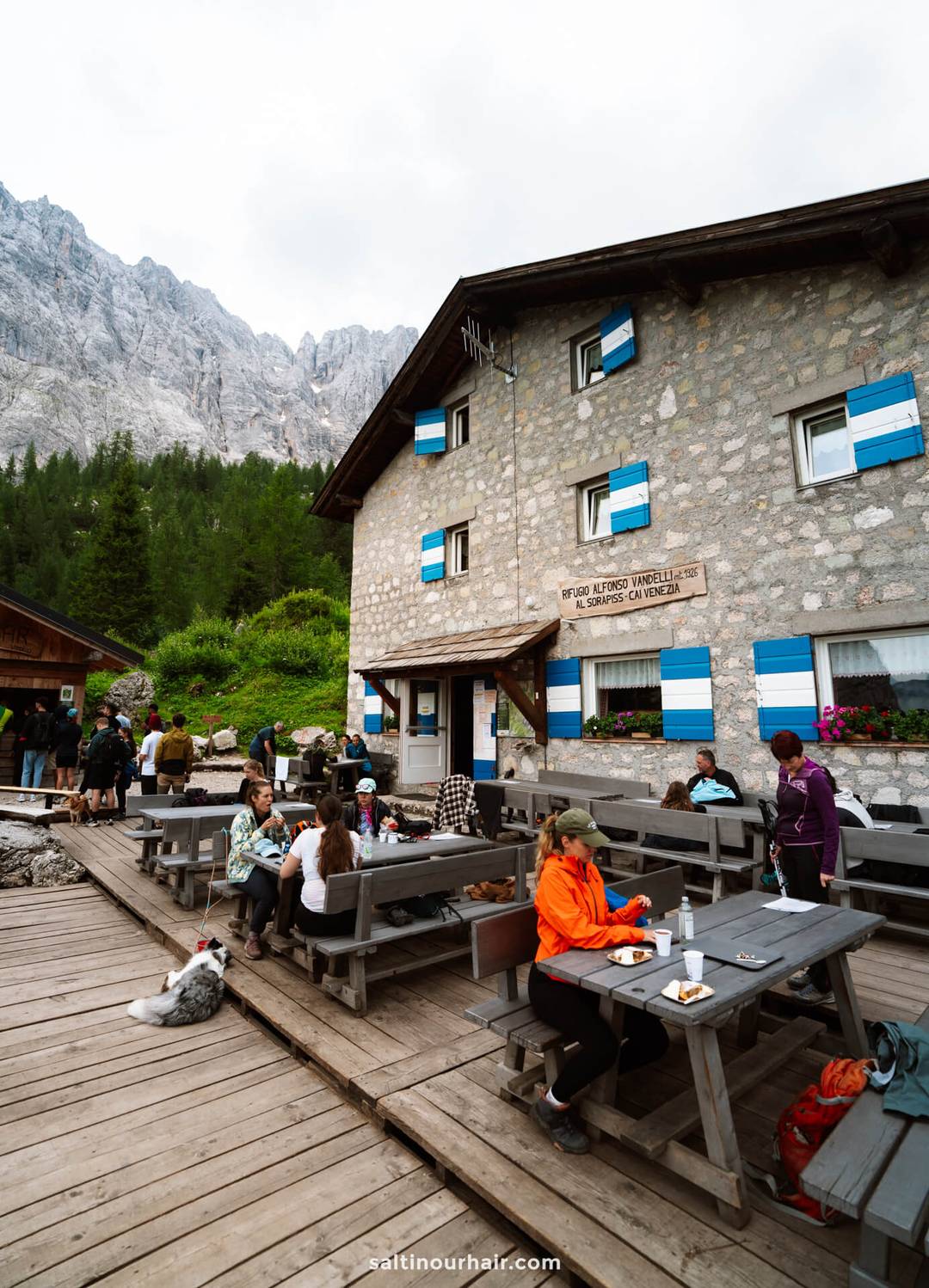 People sit and eat at outdoor picnic tables in front of Refugio Vandelli, a stone mountain lodge with blue-shuttered windows; forest and the rocky peaks of the Dolomites, near Lago di Sorapis, form a stunning backdrop.