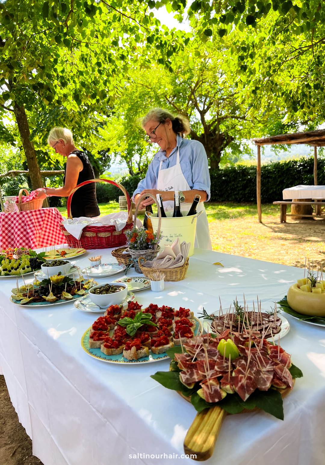 Two women prepare and arrange appetizers on outdoor tables covered with white and checkered cloths, surrounded by greenery and shaded by trees on a charming Tuscany farm Italy.