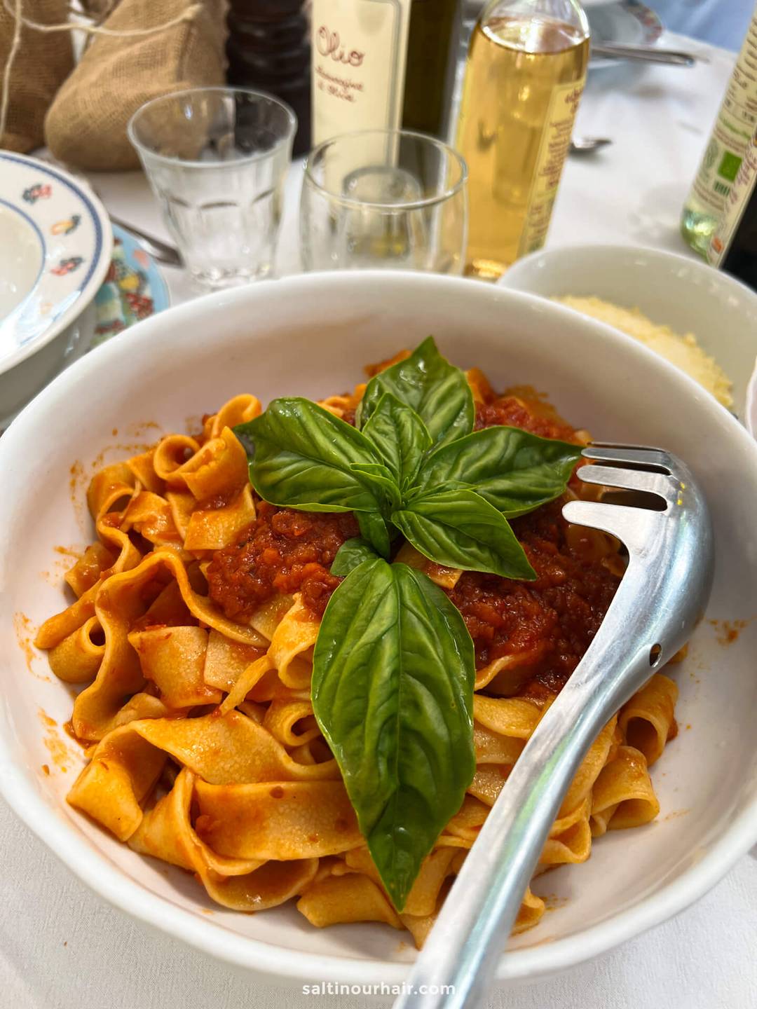A bowl of tagliatelle pasta with tomato sauce, topped with fresh basil leaves, sits on a table set with glasses, bottles, and a serving fork&mdash;a true farm to table experience at Fattoria la Vialla tuscany italy