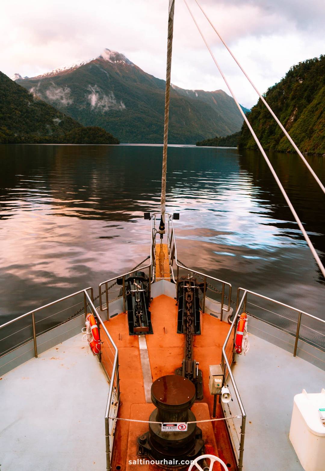 Experience the view during an overnight cruise on Doubtful Sound&mdash;a tranquil trip from Queenstown, New Zealand&mdash;surrounded by mountains, trees, and a distant snow-capped peak beneath a cloudy sky.