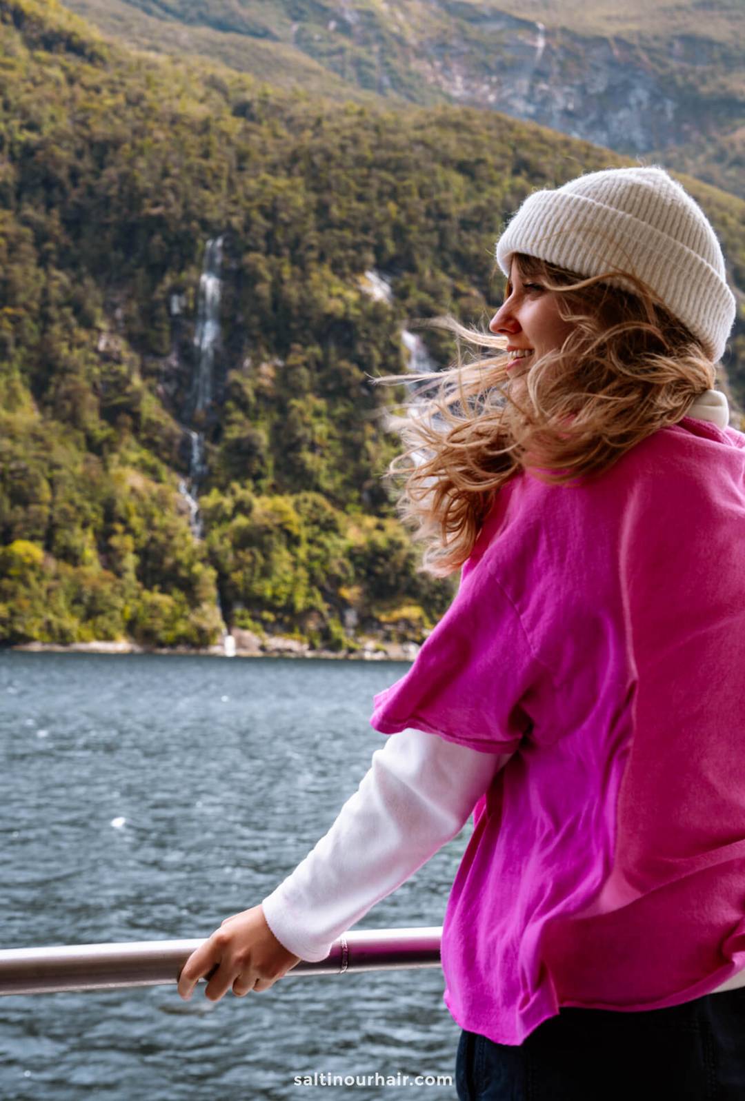 Woman in a pink shirt and white beanie stands by a railing on a boat, admiring the stunning lake and forested mountains&mdash;one of the best things to do on a Milford Sound day trip from Queenstown, New Zealand.