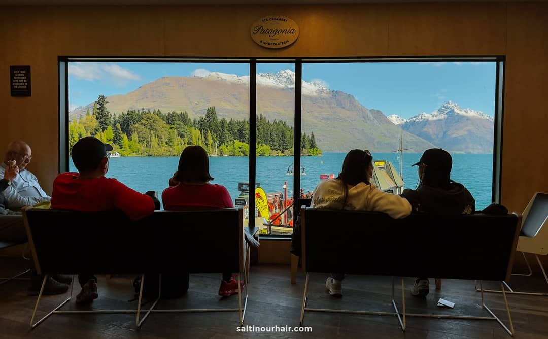 Four people sit indoors on chairs facing large windows at Patagonia Chocolates with a view of a lake, mountains, and a docked boat under a clear sky&mdash;capturing one of the best things to do in Queenstown.