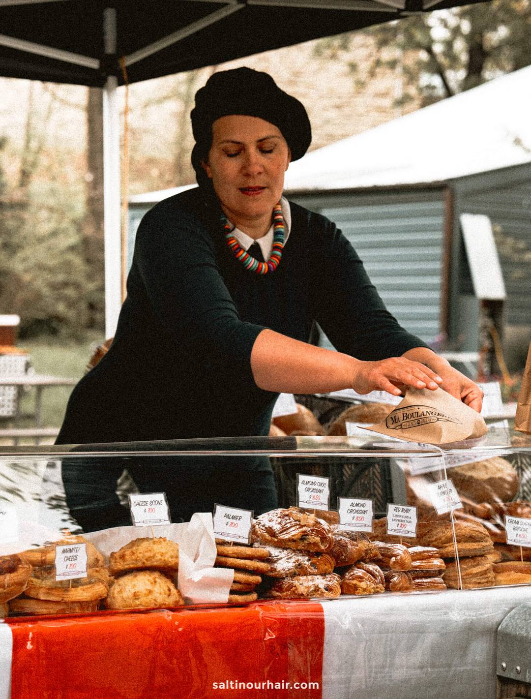 A woman in a black beret and colorful necklace arranges baked goods at an outdoor market stall on the sunday market in arrow town &mdash;one of the many things to do in Queenstown. Pastries and breads are neatly displayed with labels on the counter.