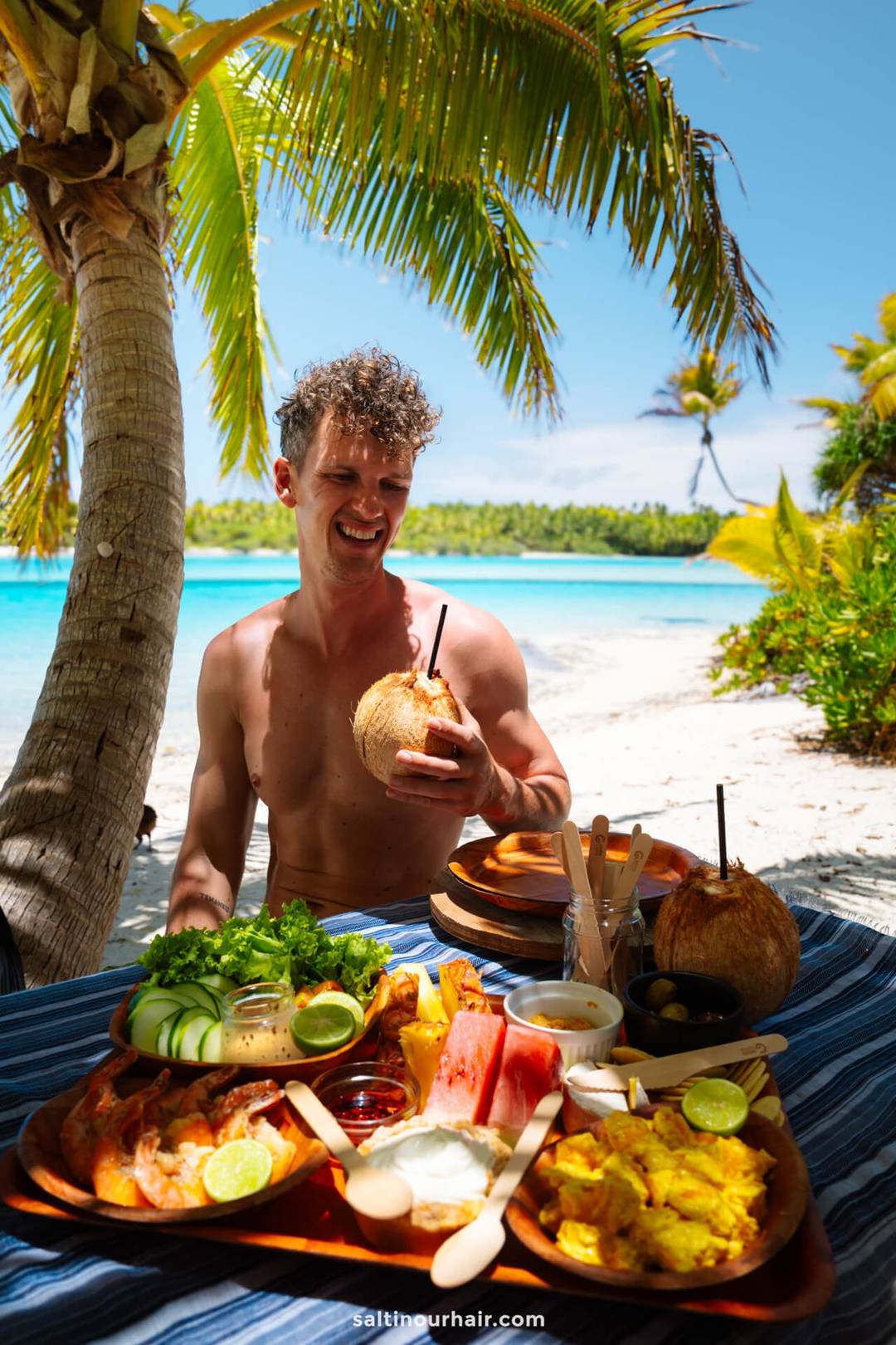 A shirtless man sits at a tropical beachside table in one foot island, aitutaki, cook islands. Holding a coconut drink, with assorted fruit and food dishes in front of them under palm trees