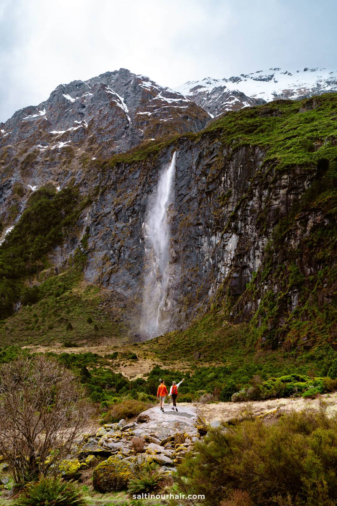 Two people in red jackets walk along the Rob Roy Track in new zealand, heading toward a tall waterfall cascading down a rocky, green mountainside topped with snow. The perfect day on a new zealand road trip