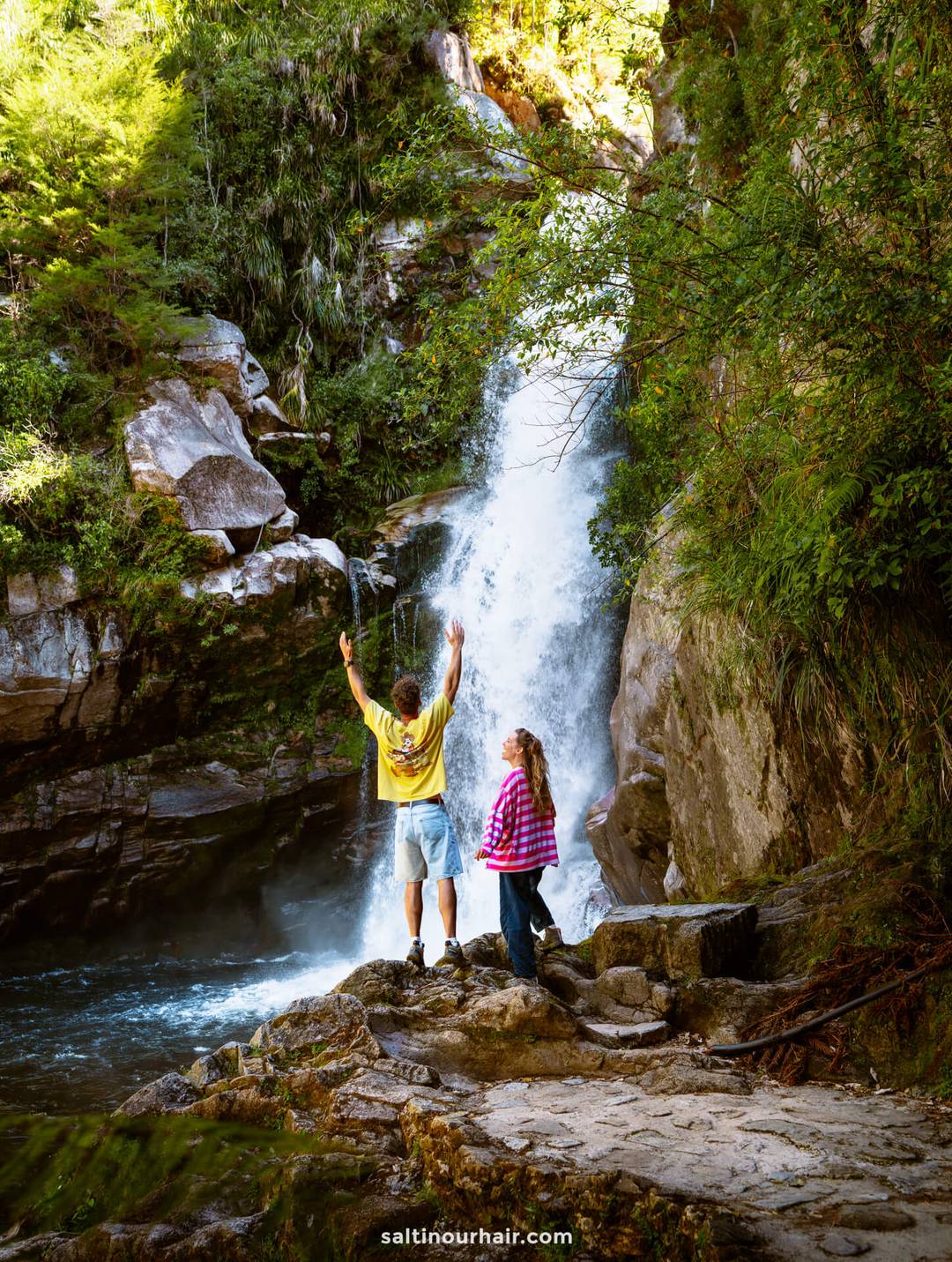 Two people stand on rocks near a waterfall in the forested Abel Tasman National Park; one raises their arms while the other gazes at the water.