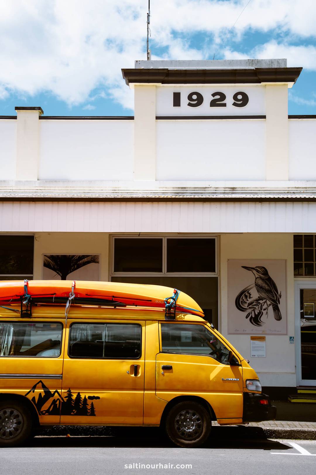 A yellow van with mountain and tree graphics is parked in front of a white building labeled 1929, with two kayaks on its roof&mdash;ready for an adventure to Abel Tasman National Park.