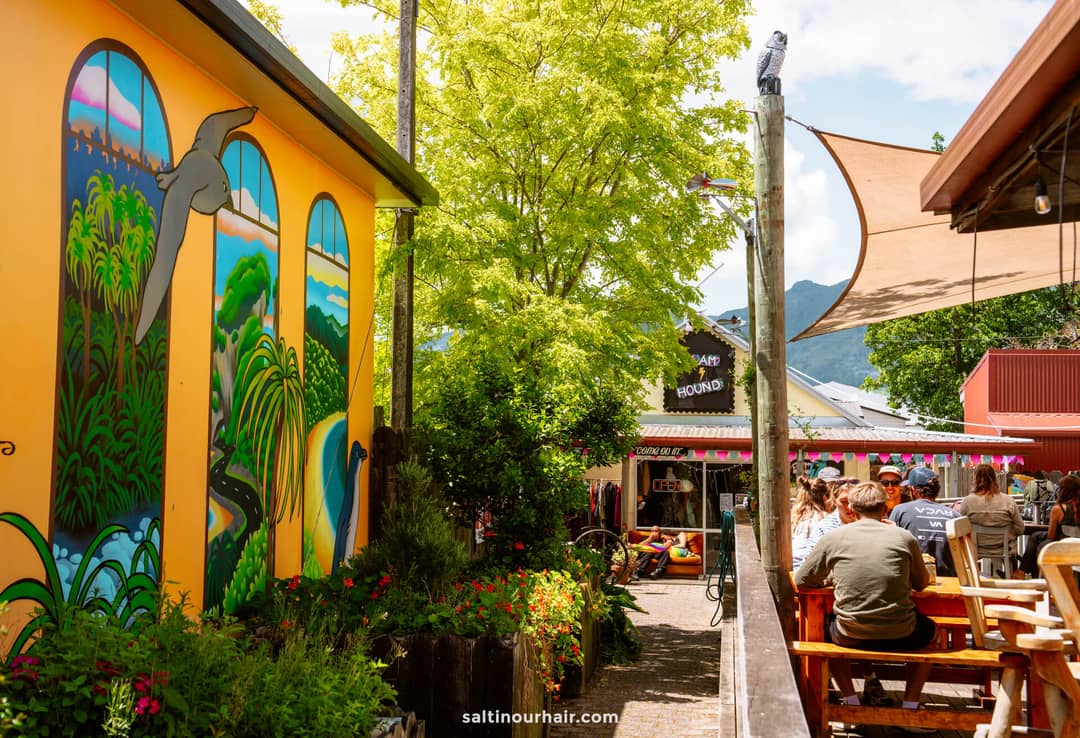 Outdoor seating area with wooden tables, people dining, a colorful mural on a yellow wall, trees, and festive bunting under a sunny sky&mdash;perfect for relaxing after exploring Abel Tasman National Park.