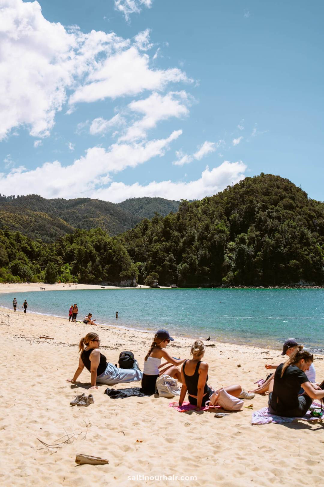 People relaxing on a sandy beach near turquoise water in Abel Tasman National Park, with forested hills and a partly cloudy sky in the background.