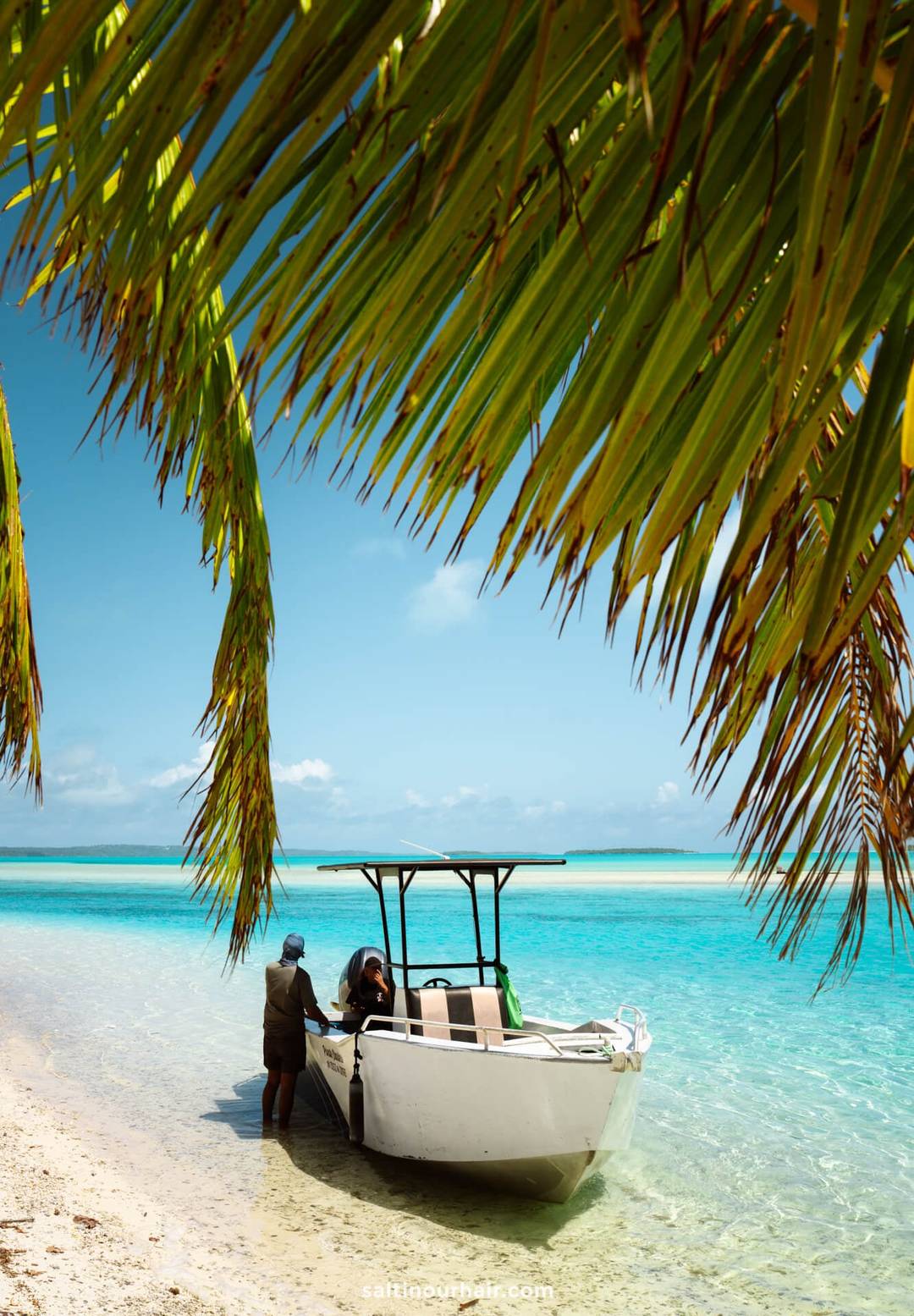 A small boat is anchored in shallow turquoise water near the sandy beach of Aitutaki island, with two people standing on the shore and palm fronds hanging overhead.