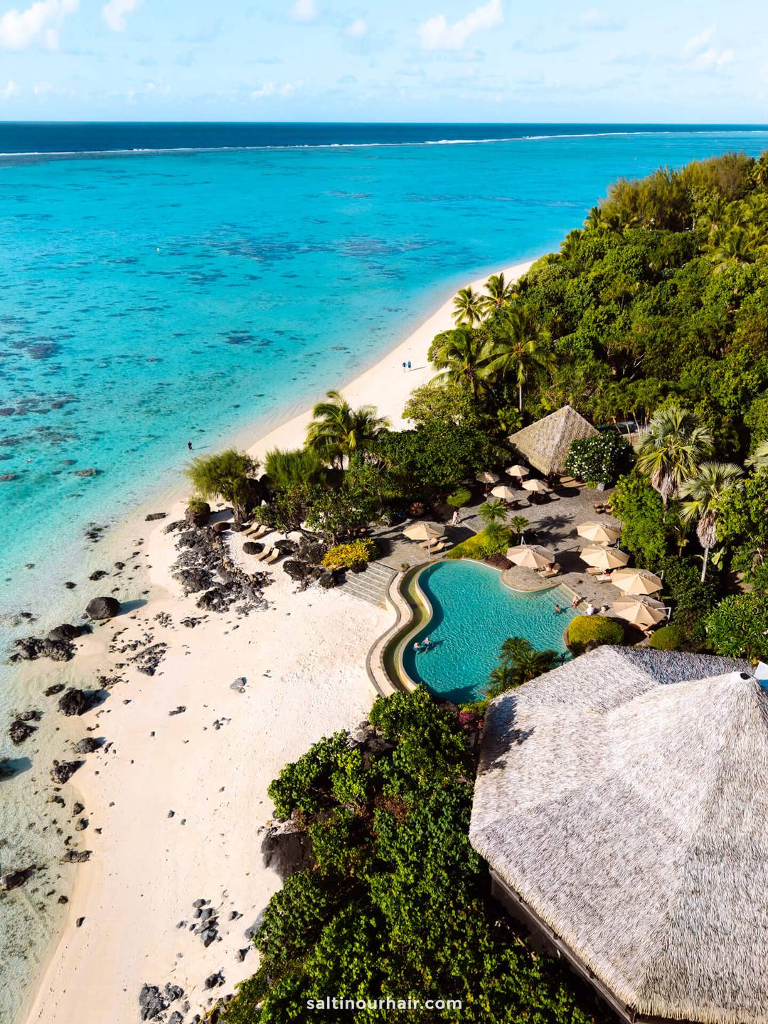 Aerial view of pacific beach resort aitutaki with a pool, palm trees, sandy beach, and clear turquoise ocean water in cook islands