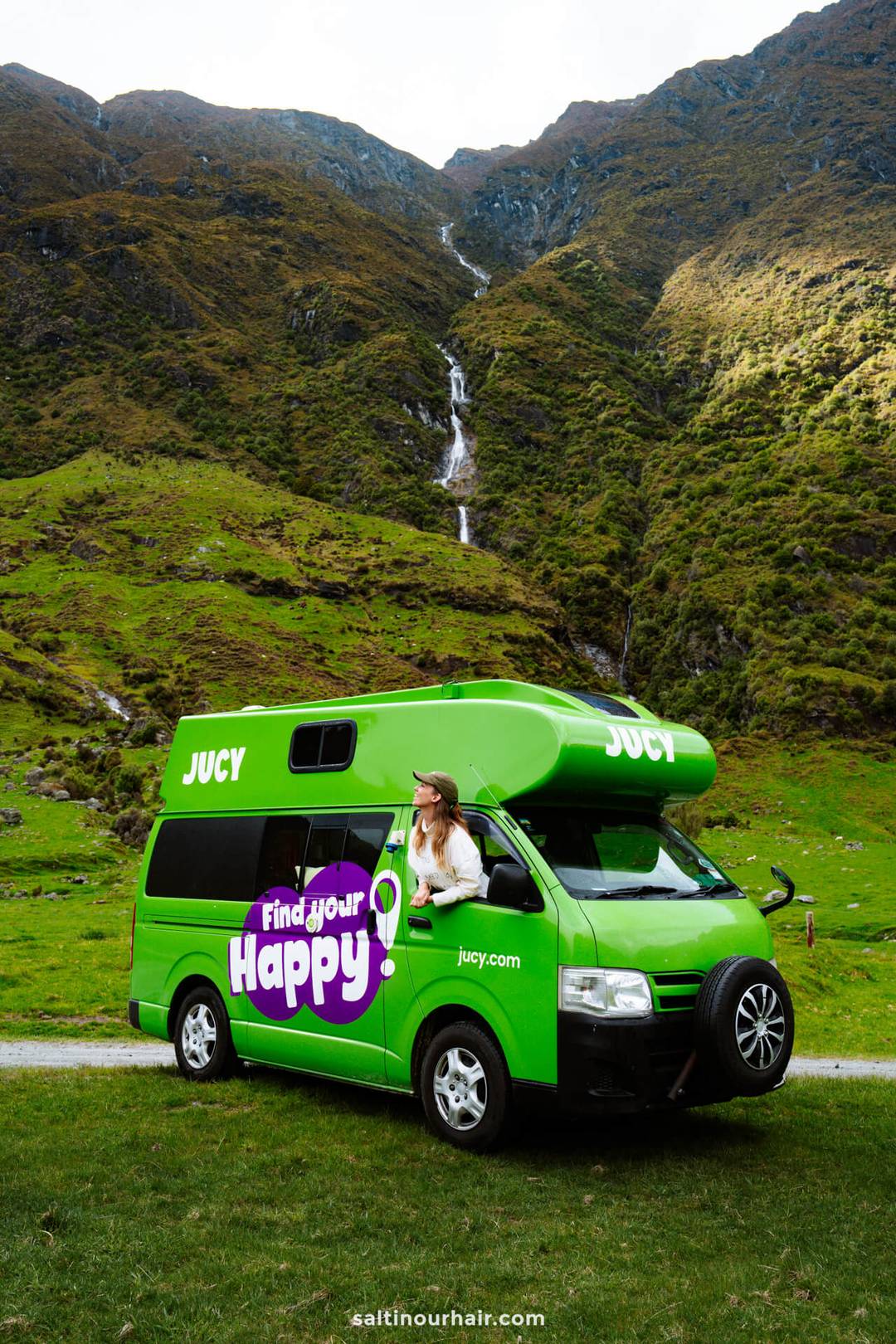 A woman in a hat leans out of the window of a bright green camper van parked on grass along the scenic Rob Roy Track, with mountains and a waterfall in the background.