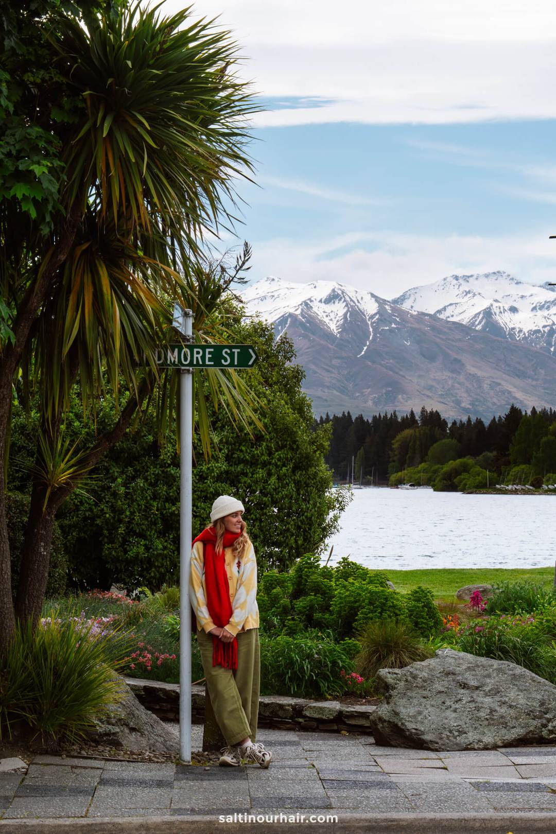a woman leaning against a street sign in wanaka new zealand with snow capped mountains and a lake in the background