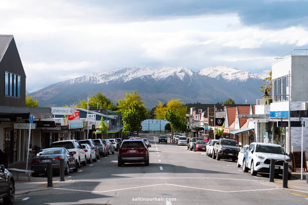 A quiet street in Wanaka, New zealand, lined with parked cars and shops, with snow-capped mountains