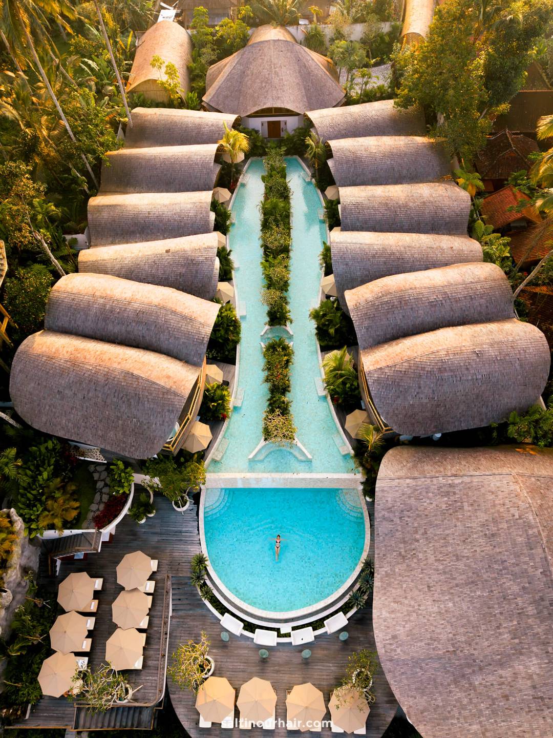 Aerial view of a bambootel tropical resort with thatched-roof villas surrounding a long, narrow pool leading to a round pool where a person is swimming; lounge chairs and umbrellas are nearby.