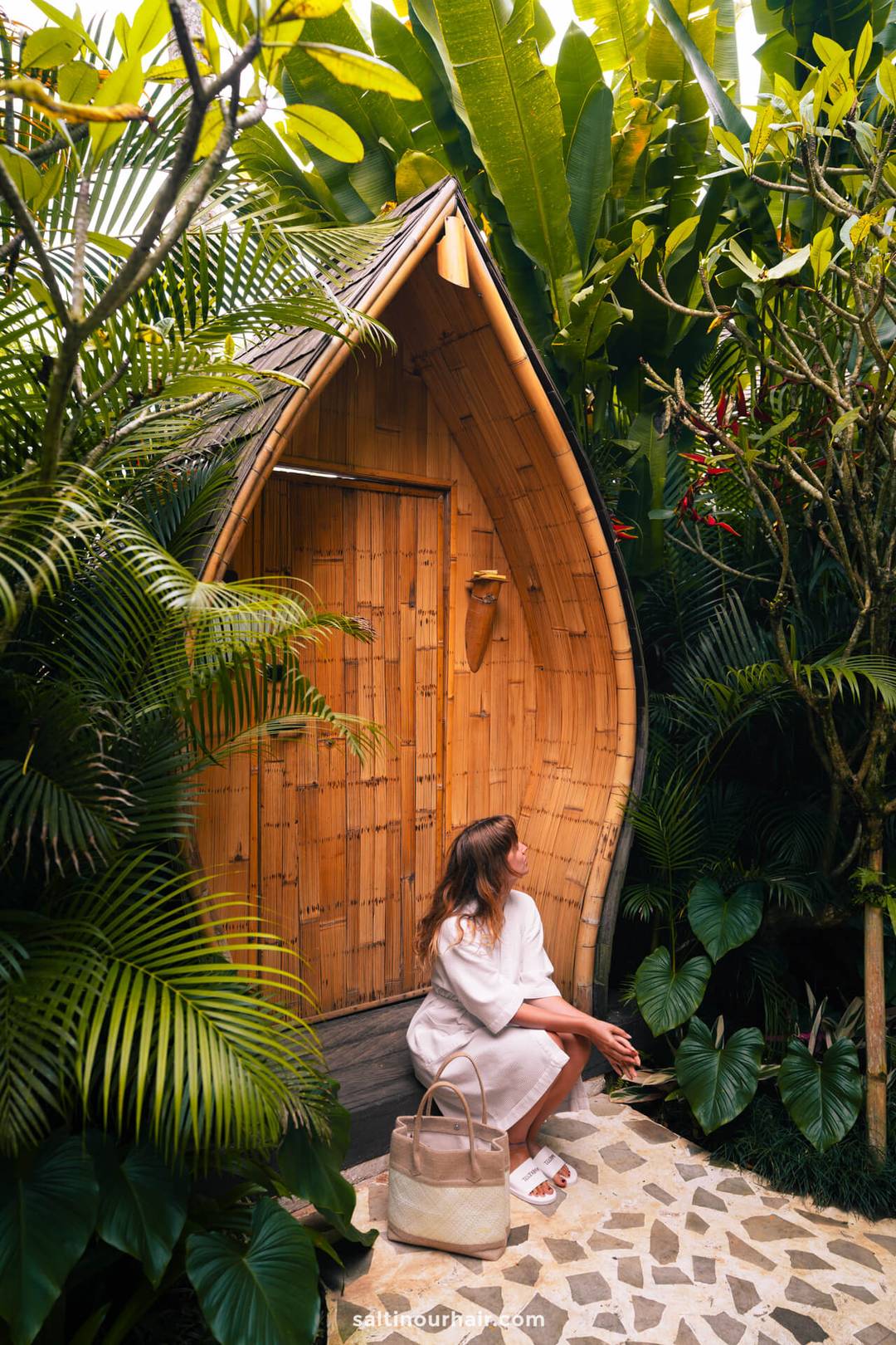 A woman in a white dress sits on the steps of a charming bambootel, surrounded by lush tropical plants, holding sunglasses and a beige bag.