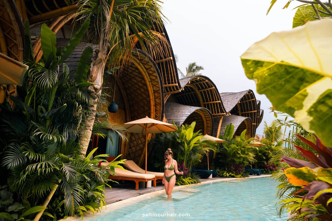 A woman in a green bikini walks by a pool surrounded by tropical plants and bambootel villas with elegant, curved roofs.
