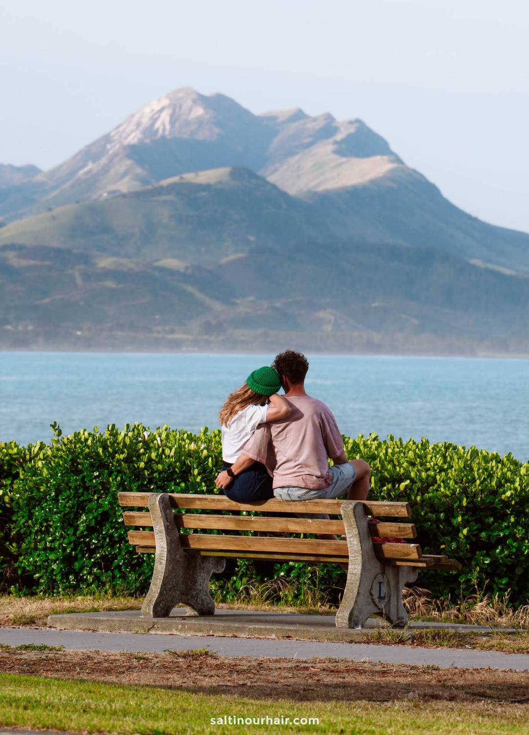 A couple sits on a wooden bench facing a body of water, with mountains and the Kaikōura Seal Colony in the background.
