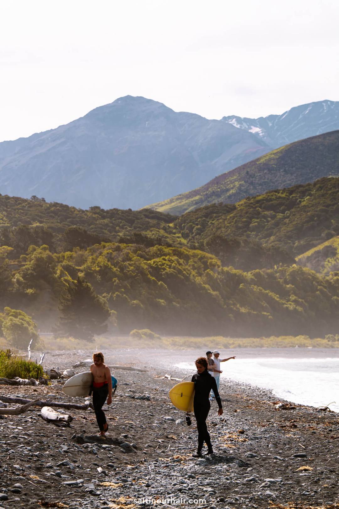 Three people carrying surfboards walk along a rocky beach near the Kaikōura Seal Colony, with mountains and lush greenery in the background.