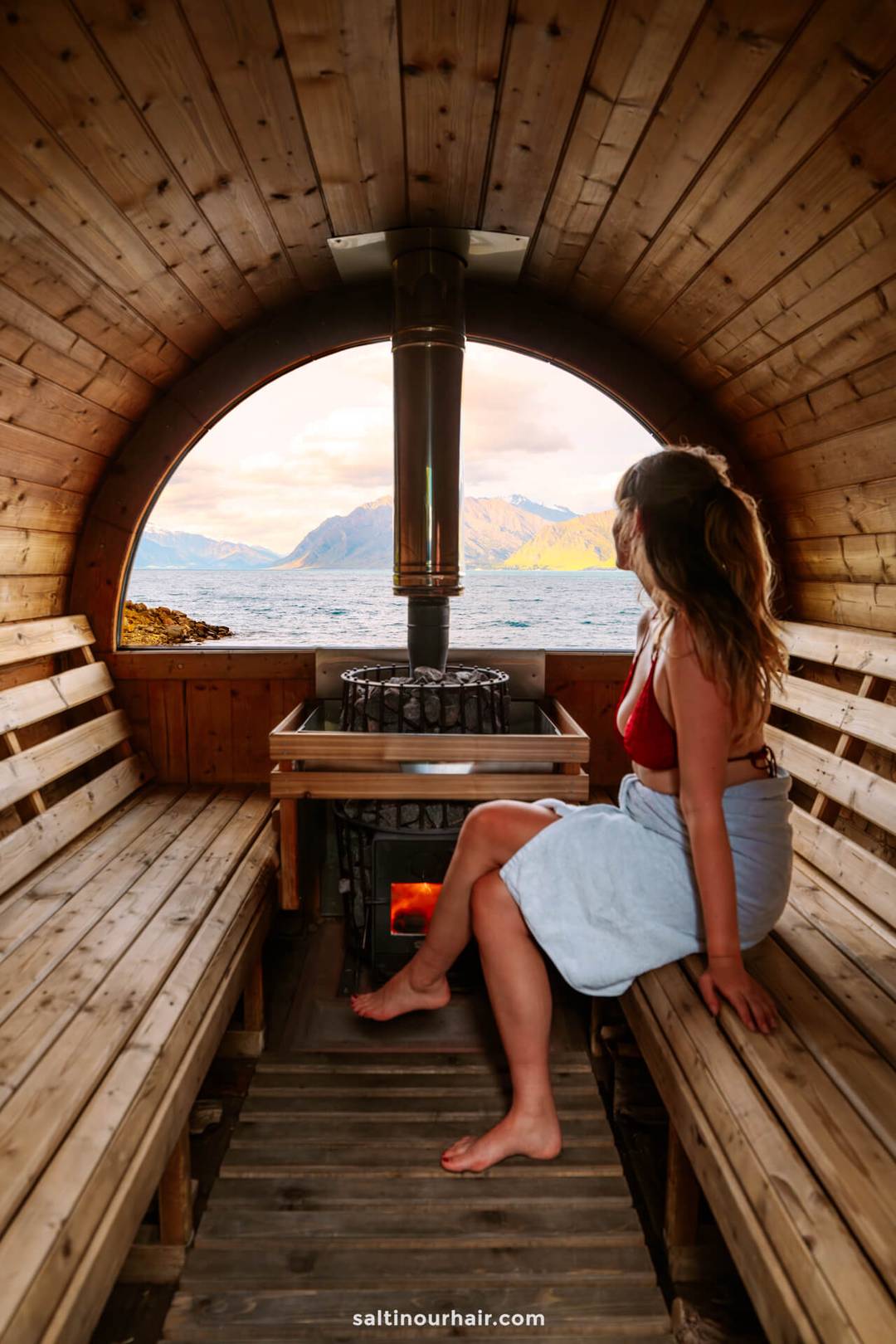 A woman in a towel sits inside a wooden sauna in Wānaka, gazing out at mountains and water through a large circular window at the end of the sauna.