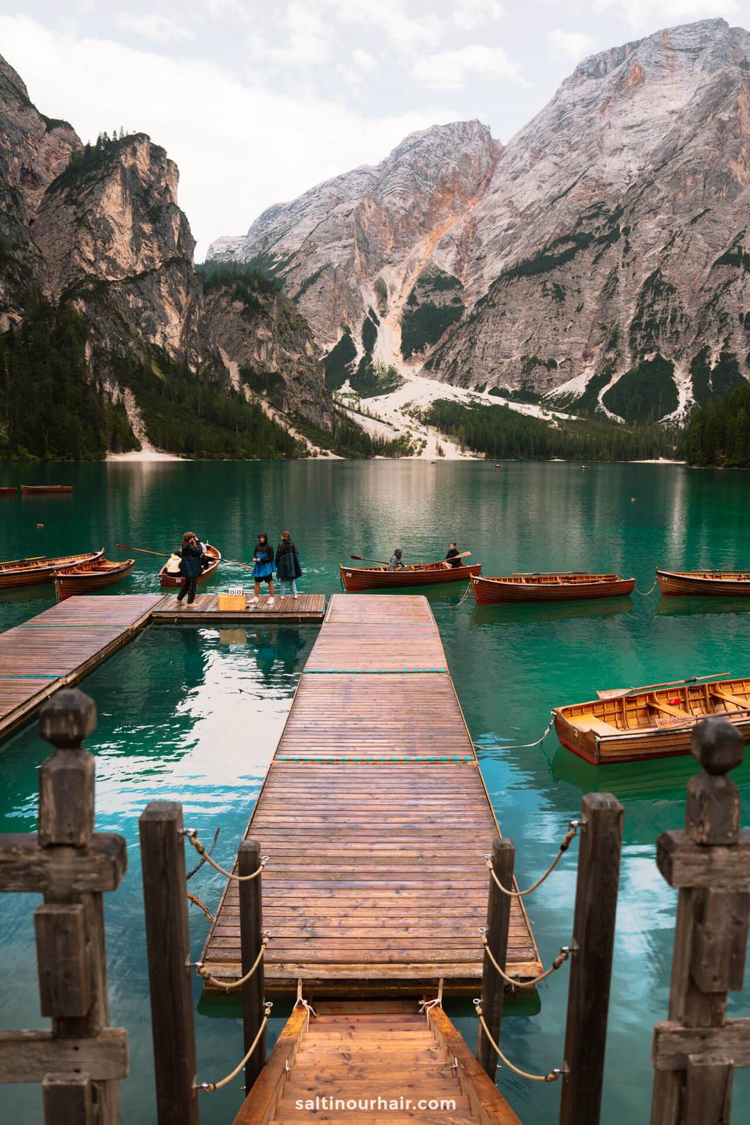 A wooden dock extends into a turquoise lake with several wooden boats, surrounded by mountains and forested slopes under a cloudy sky-an idyllic spot to include in your Dolomites itinerary.