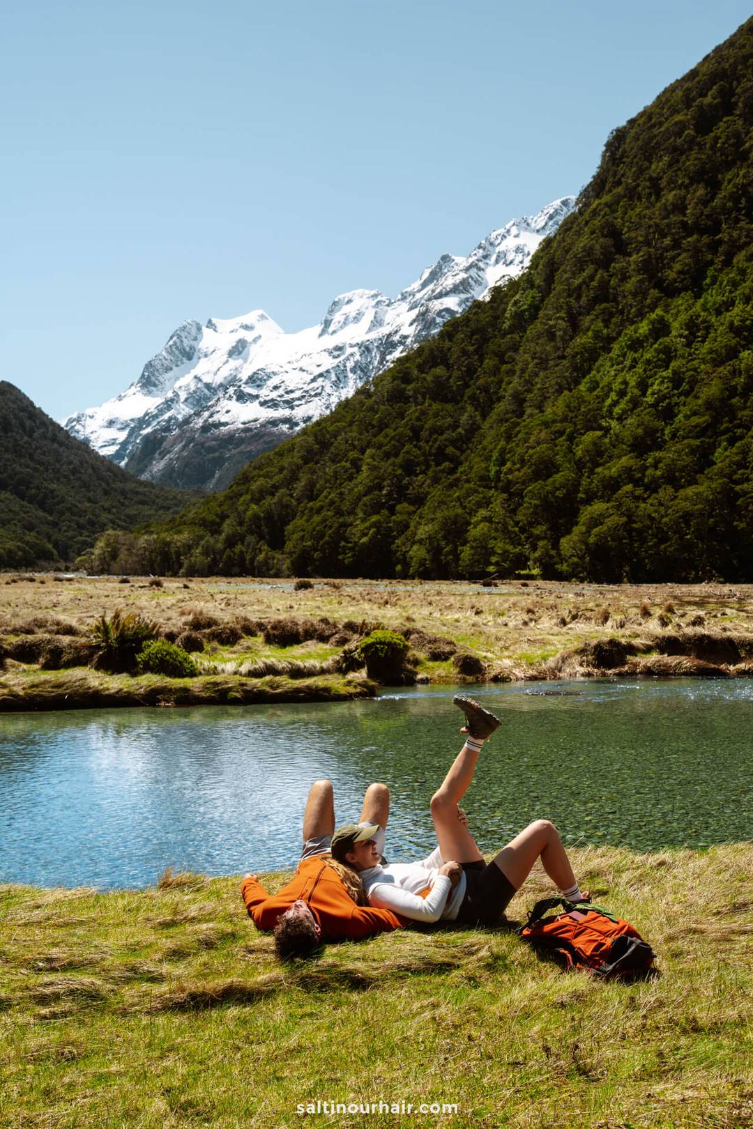 Two people lie on the grass by a clear river, surrounded by green hills and snow-capped mountains under a clear blue sky&mdash;an idyllic scene straight from a New Zealand South Island itinerary.