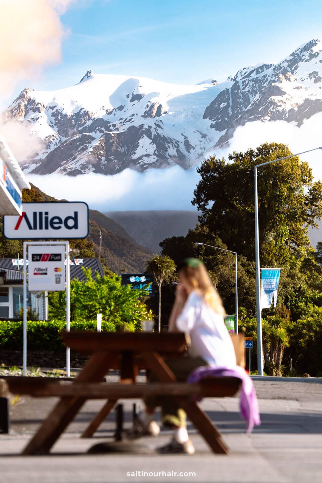 A person sits at a picnic table near an Allied gas station with snow-covered mountains and a blue sky in the background&mdash;a classic stop on any New Zealand South Island itinerary.