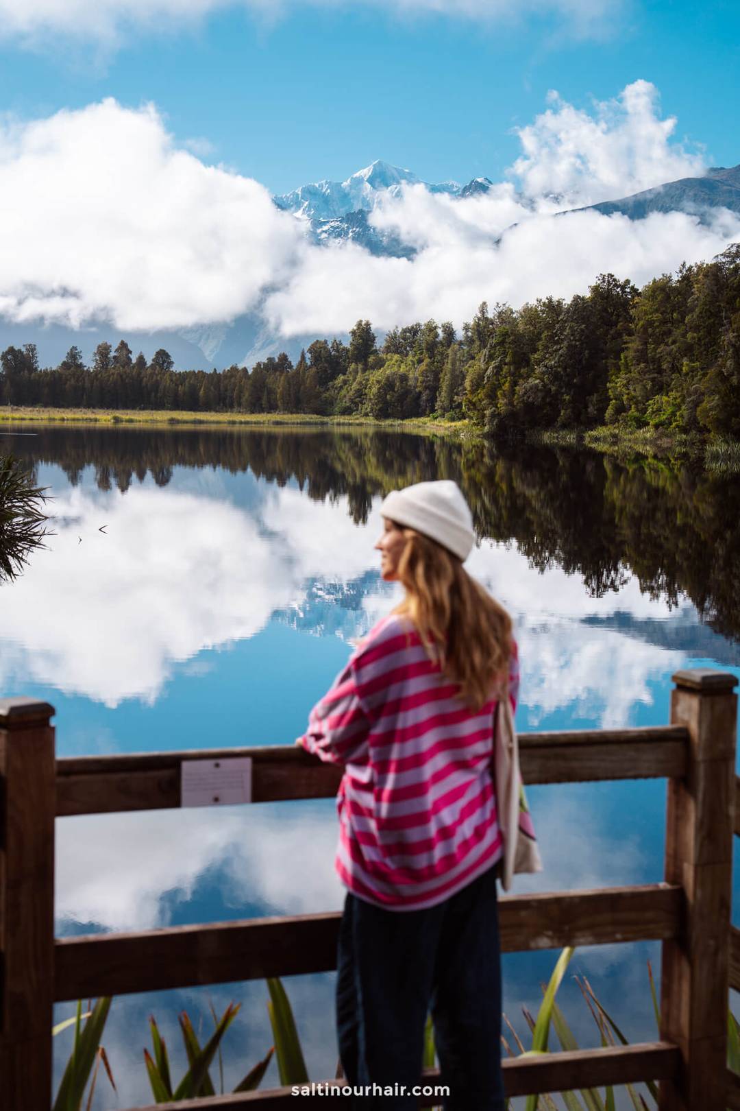 Person in a pink striped sweater and white beanie stands at a wooden railing, overlooking the calm lake matheson with forest and snow-capped mountains&mdash;a scene straight from a New Zealand South Island itinerary.