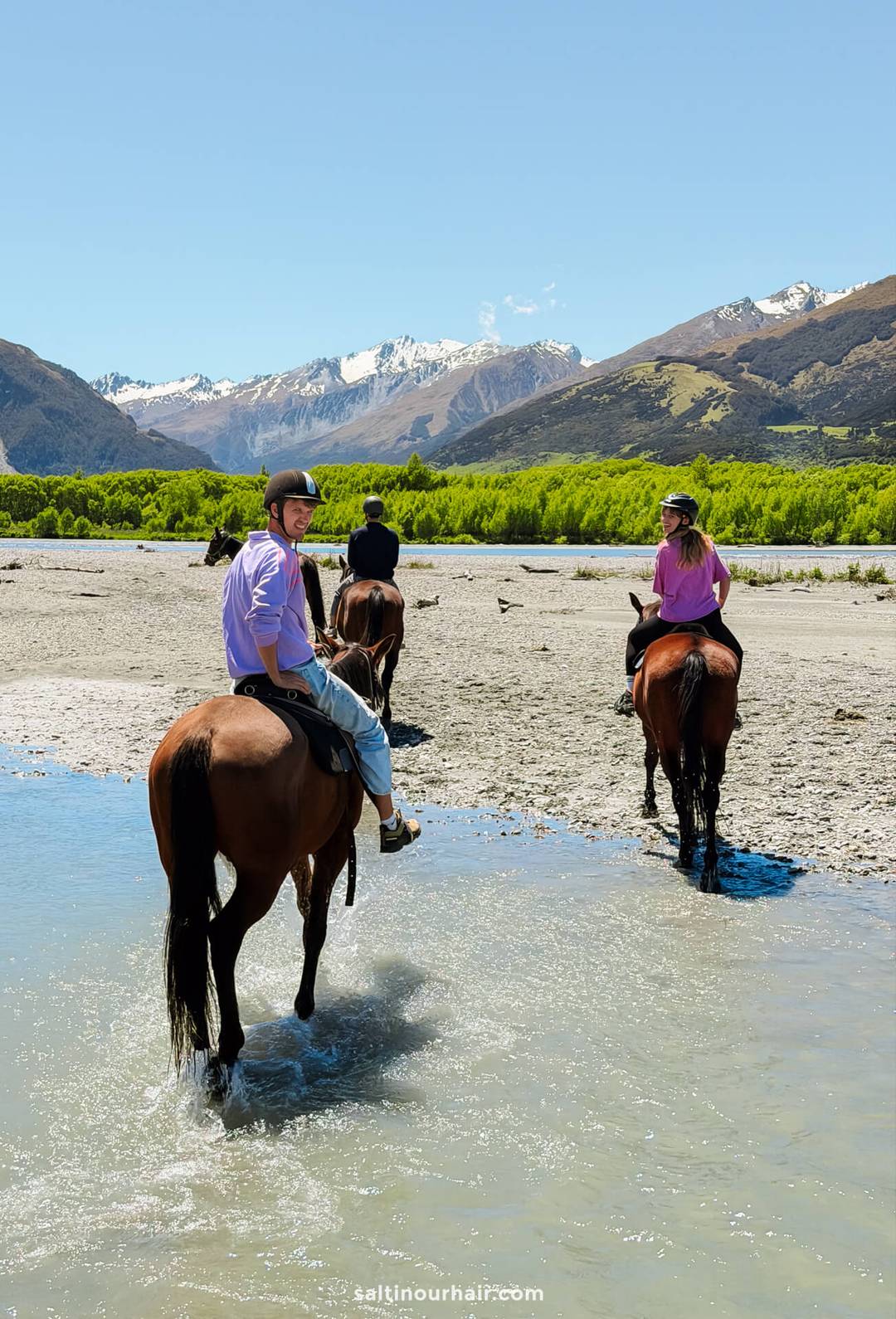 Three people on horseback cross a shallow river surrounded by mountains and greenery&mdash;a scene straight from a New Zealand South Island itinerary&mdash;on a clear, sunny day.