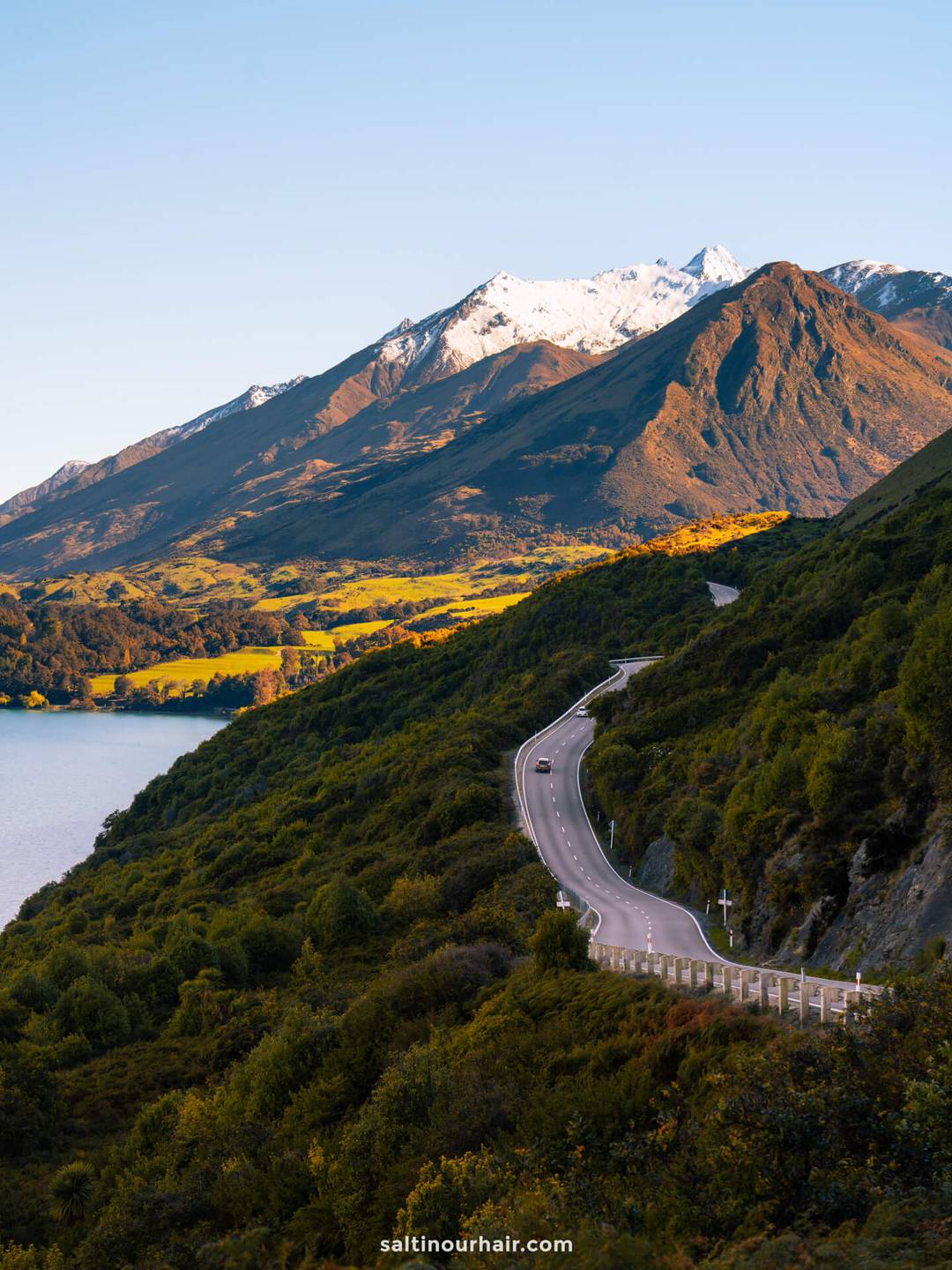 A winding road passes through green hills with a lake on the left and snow-capped mountains in the background&mdash;an idyllic stop on any New Zealand South Island itinerary under a clear sky.