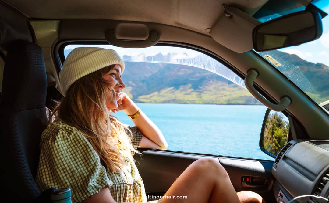 A woman wearing a white beanie sits in the passenger seat of a vehicle, looking out at a lake and mountains&mdash;an inspiring moment on her New Zealand South Island itinerary.