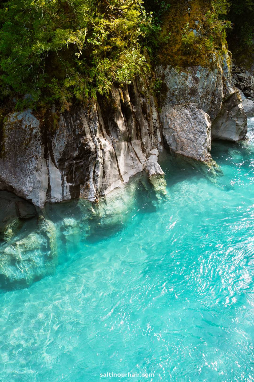 Clear turquoise river water of the Blue Pools flows beside rocky cliffs with green foliage above&mdash;a perfect highlight for any New Zealand South Island itinerary. Sunlight reflects on the water&rsquo;s surface, creating a serene and inviting scene.