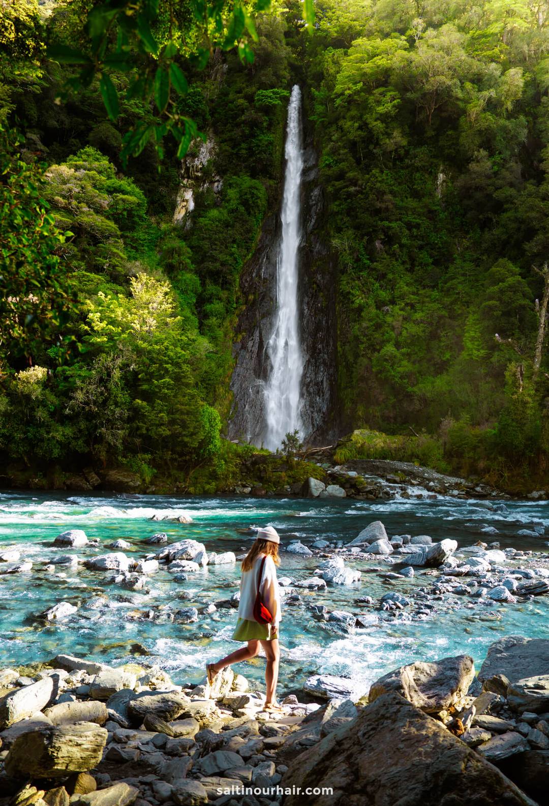 A person wearing a hat and holding a bag stands by a rocky riverbank with the tall Thunder Creek Falls and dense green forest in the background&mdash;an ideal scene for any New Zealand South Island itinerary.