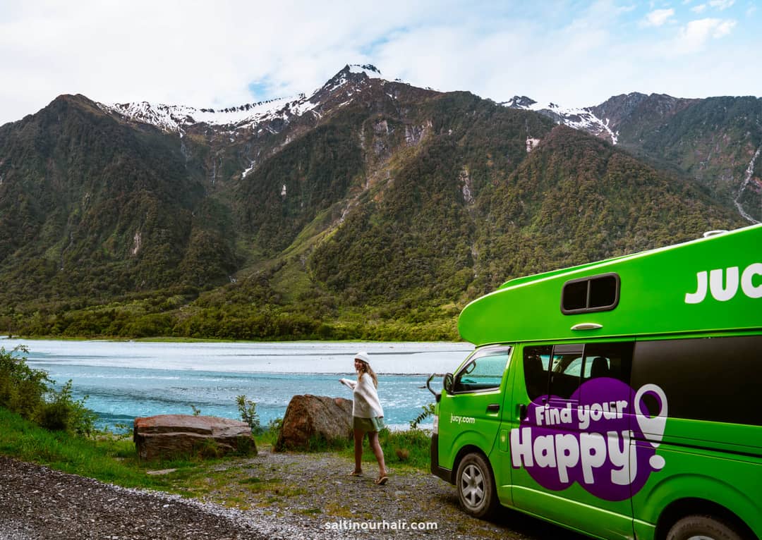 A green campervan parked by a lake with mountains in the background; a person stands near the water holding a phone, perhaps mapping out their New Zealand South Island itinerary.