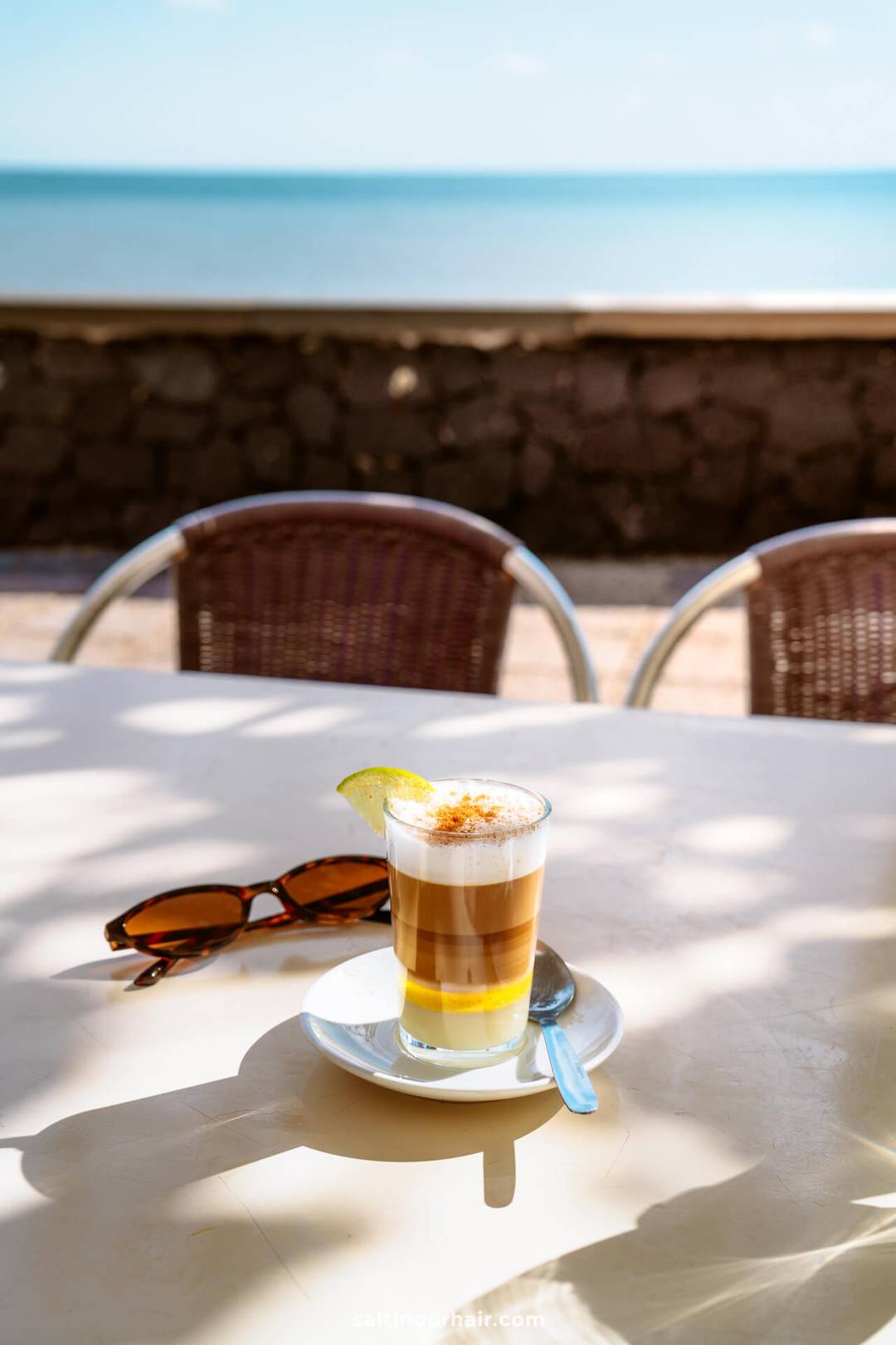 A layered coffee with a lemon slice sits on a saucer with a spoon on a sunlit outdoor table in La Palma, next to sunglasses, with the ocean and a stone wall in the background&mdash;a perfect scene for any Canary Islands travel guide.