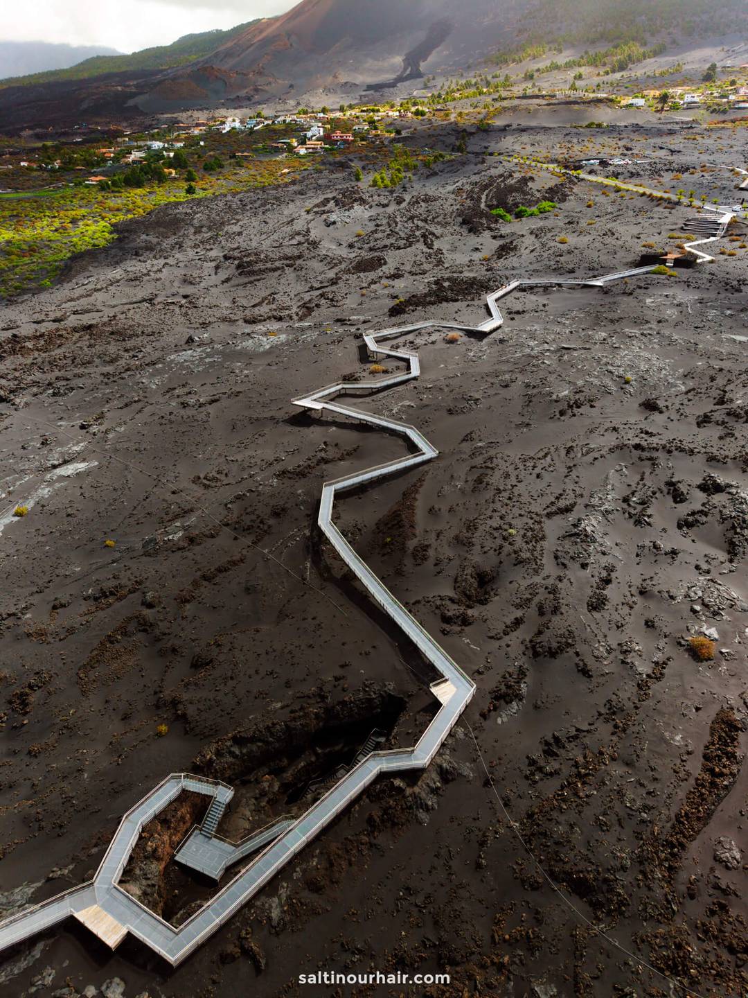A zigzag wooden walkway crosses a barren, volcanic ash landscape on La Palma, with small houses and green vegetation in the background&mdash;an iconic sight in this Canary Islands travel guide.