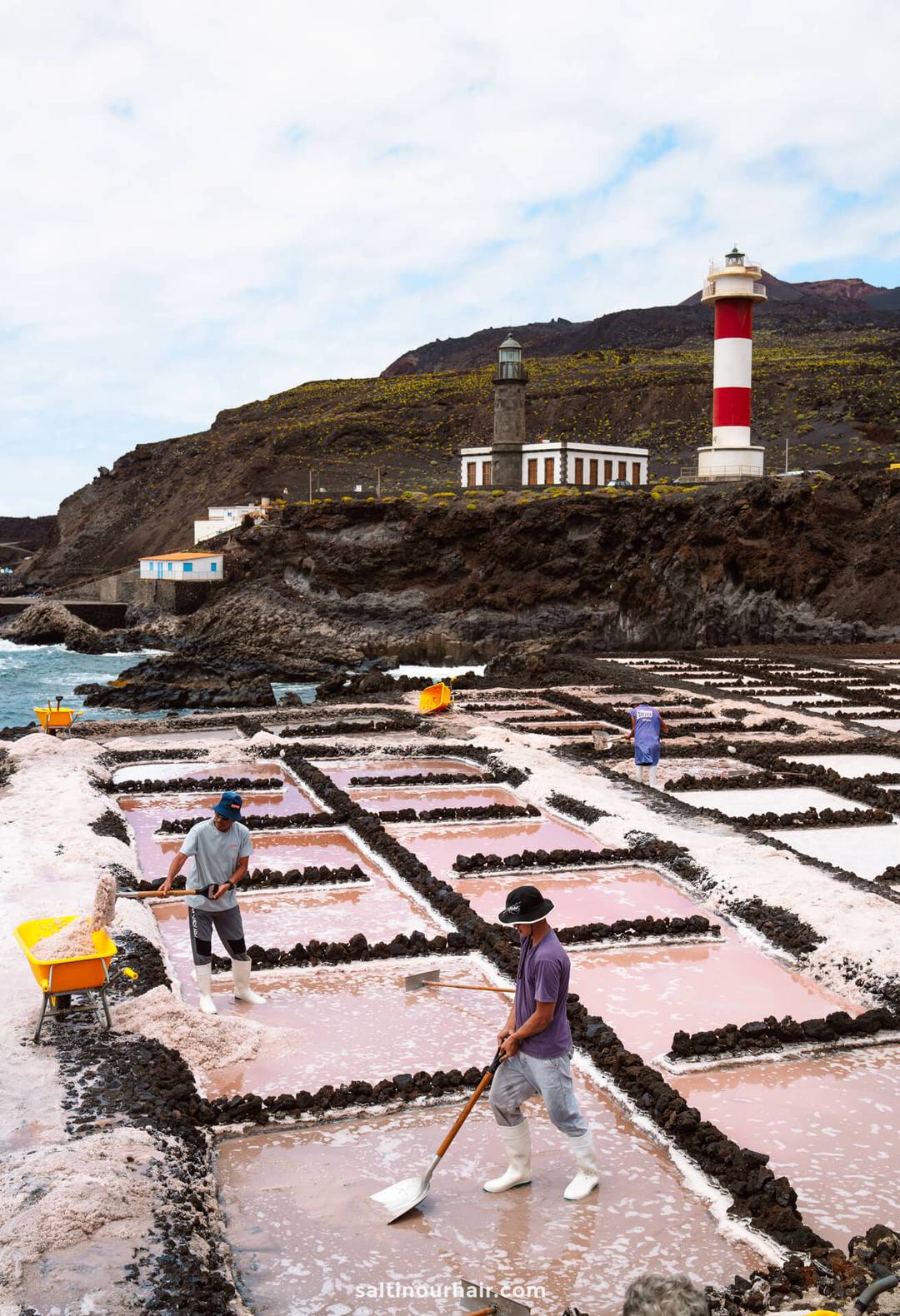 Workers harvest salt from rectangular salt pans near the coast of La Palma, with a lighthouse and buildings on a rocky hill in the background&mdash;a striking scene in the Canary Islands worthy of any travel guide.
